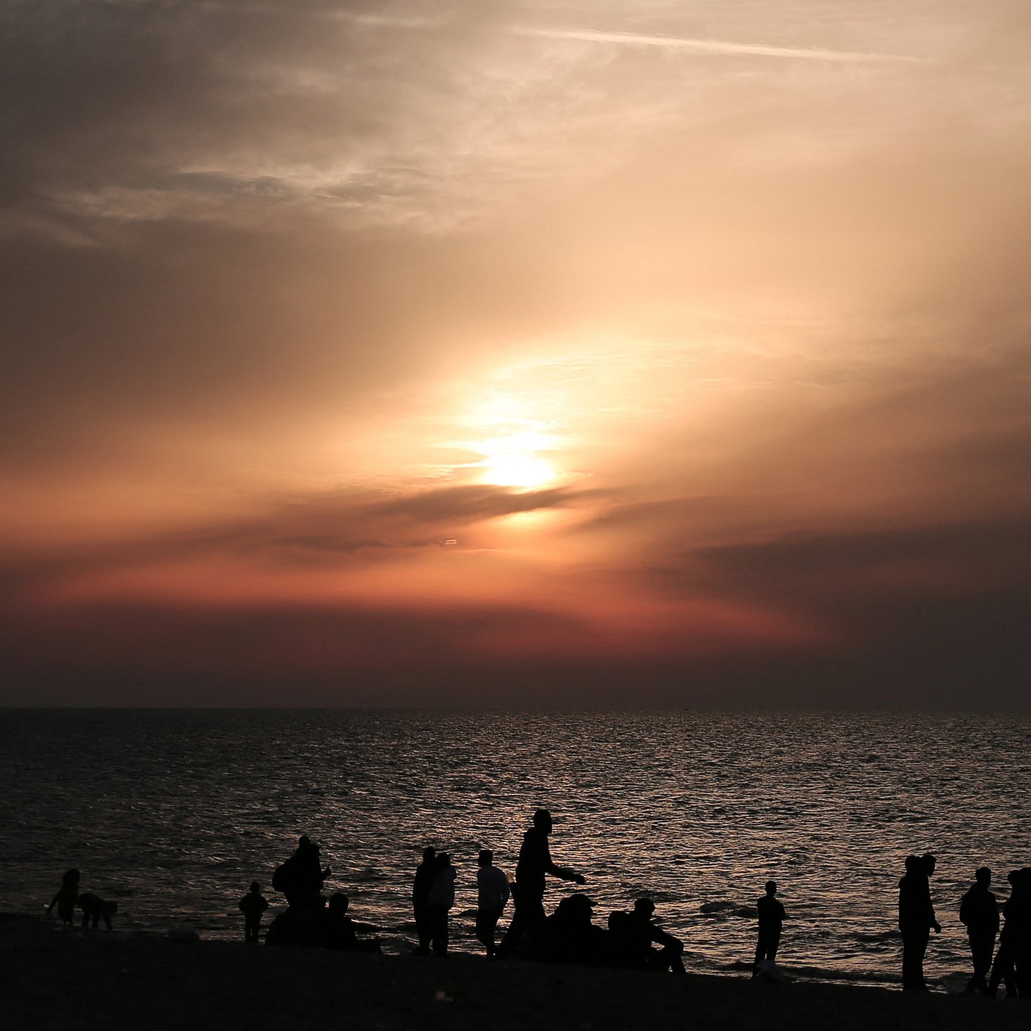L'image montre une plage au crépuscule, avec des nuages qui tamisent la lumière du soleil couchant. De nombreuses silhouettes de personnes se déplacent le long du rivage, profitant de la scène. La mer reflète les couleurs douces du ciel, créant une atmosphère tranquille et paisible. Les silhouettes des individus ajoutent une dynamique à l'image, suggérant une ambiance conviviale et joyeuse.