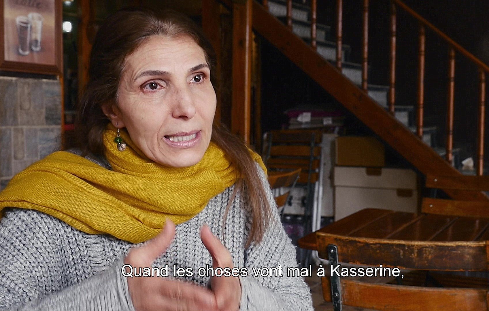 L'image montre une femme en train de parler, assise dans un environnement intérieur. Elle porte un vêtement gris avec un foulard jaune autour du cou. L'arrière-plan présente des escaliers en bois et des tables en bois, ce qui donne une ambiance chaleureuse et intime. Elle semble s'exprimer avec passion, évoquant probablement des enjeux importants liés à la ville de Kasserine.