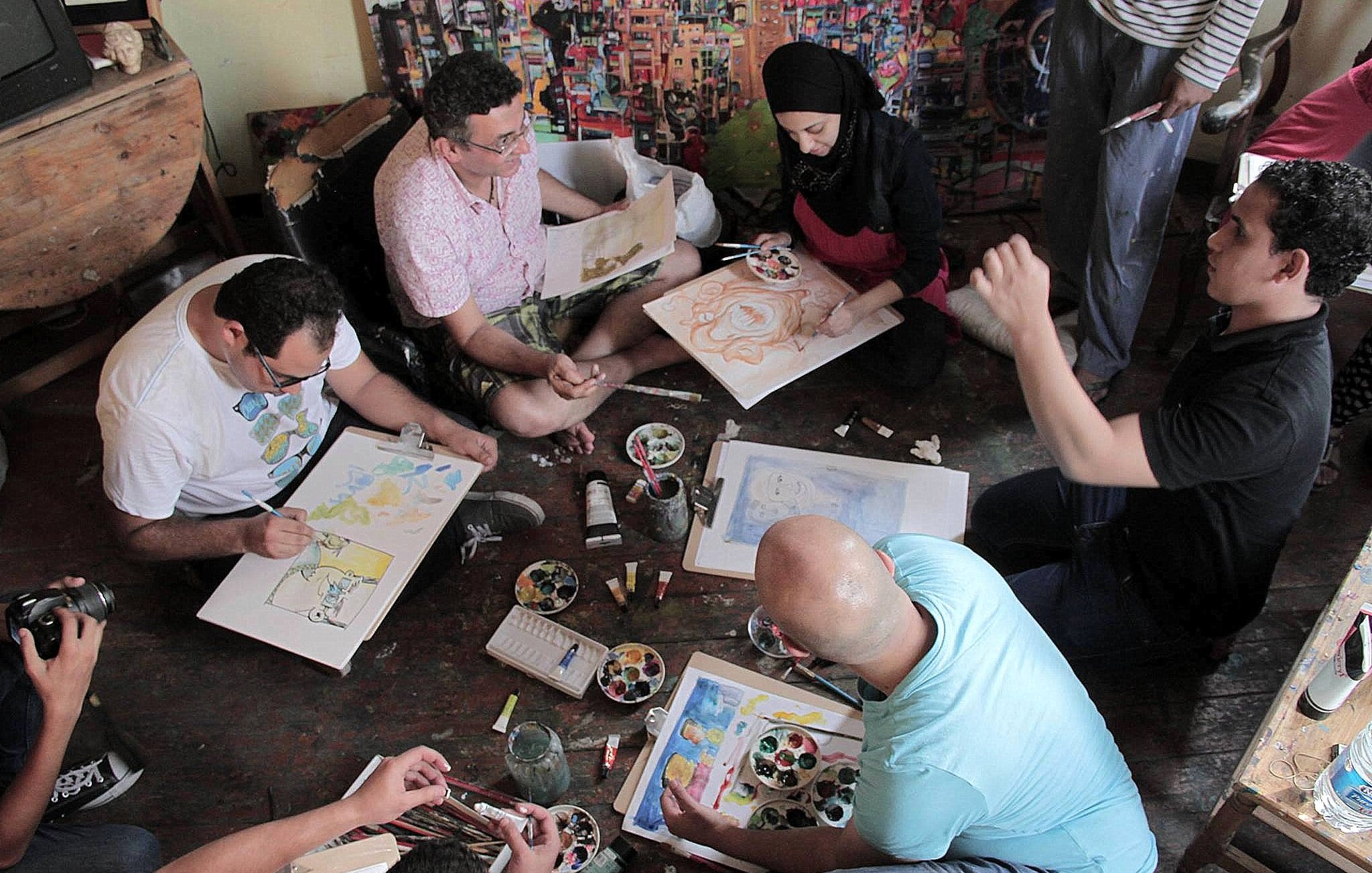 L'image montre un groupe de personnes assises sur le sol dans un espace créatif. Elles semblent engagées dans une activité artistique, utilisant des pinceaux et de la peinture sur des feuilles de papier. Certains participants semblent discuter ou partager des idées, tandis que d'autres se concentrent sur leur travail. L'environnement est coloré, avec des œuvres d'art visibles sur les murs et une palette de peinture au centre. L'ambiance paraît conviviale et collaborative.