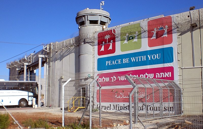 L'image montre un mur imposant, probablement un mur de séparation, avec une tour de surveillance au sommet. À la base du mur, il y a une grande pancarte qui affiche des messages liés à la paix, écrits en plusieurs langues. L'environnement est urbain, et on peut voir un bus stationné à proximité. Le tout est entouré d'une clôture barbelée, ce qui donne une impression de sécurité renforcée. Le ciel est dégagé et ensoleillé.