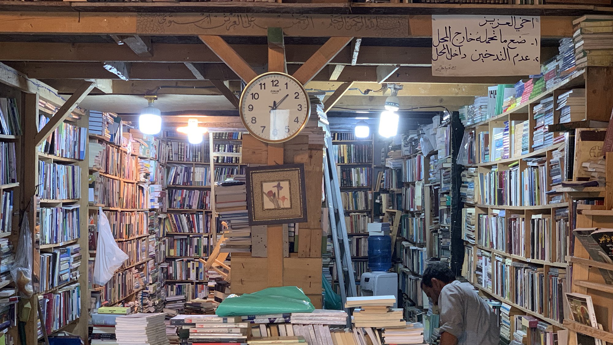 تظهر الصورة مشهداً داخلياً لمكتبة قديمة. تتراص الكتب على الرفوف الخشبية بشكل كثيف، مع ظهور بعض الكتب المكدسة على الأرض. في منتصف الصورة، يوجد ساعة حائط تشير إلى الوقت. كما يوجد شخص في الجهة اليمنى من الصورة، يبدو أنه يتفحص الكتب. الأضواء تتدلى من السقف، مما يضفي جواً دافئاً على المكان. في الأعلى، توجد لافتة مكتوبة توضح معاني تتعلق بالمكتبة.