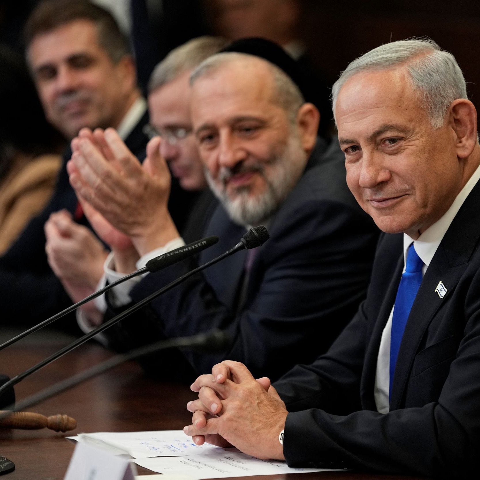 L'immagine mostra un gruppo di uomini seduti attorno a un tavolo, con una atmosfera di discussione o incontro. In primo piano, uno degli uomini, con un abito scuro e una cravatta blu, sorride e sembra ascoltare attentamente. Gli altri uomini, che si trovano accanto a lui, applaudono e sembrano essere coinvolti nella conversazione. Lo sfondo presenta un ambiente formale, probabilmente un ufficio o una sala riunioni.