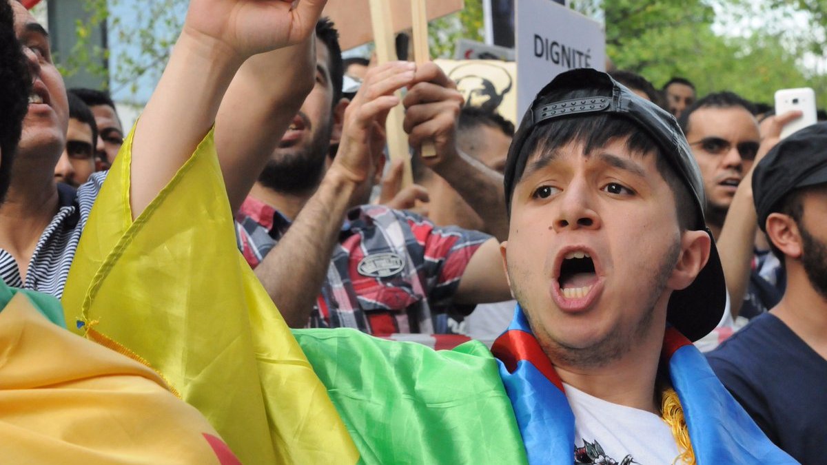 L'image montre une scène de manifestation, où un jeune homme, portant un drapeau coloré, crie avec passion. Il semble mobilisé et engagé dans la cause pour laquelle les manifestants se sont rassemblés. En arrière-plan, on peut voir d'autres personnes tenant des pancartes et exprimant leurs préoccupations. L'atmosphère est remplie d'énergie et de détermination.
