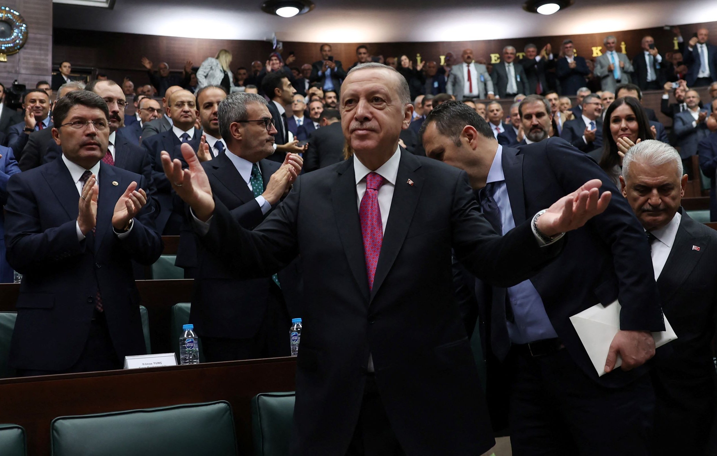 L'image montre un homme en costume, les bras écartés, se tenant au centre d'une assemblée constituée de nombreuses personnes applaudissant autour de lui. Il semble apprécier l'attention du public, dans un environnement de type parlementaire, où les membres sont assis sur des bancs et affichent des expressions variées. L'atmosphère est dynamique, suggérant un moment de célébration ou de discours important.