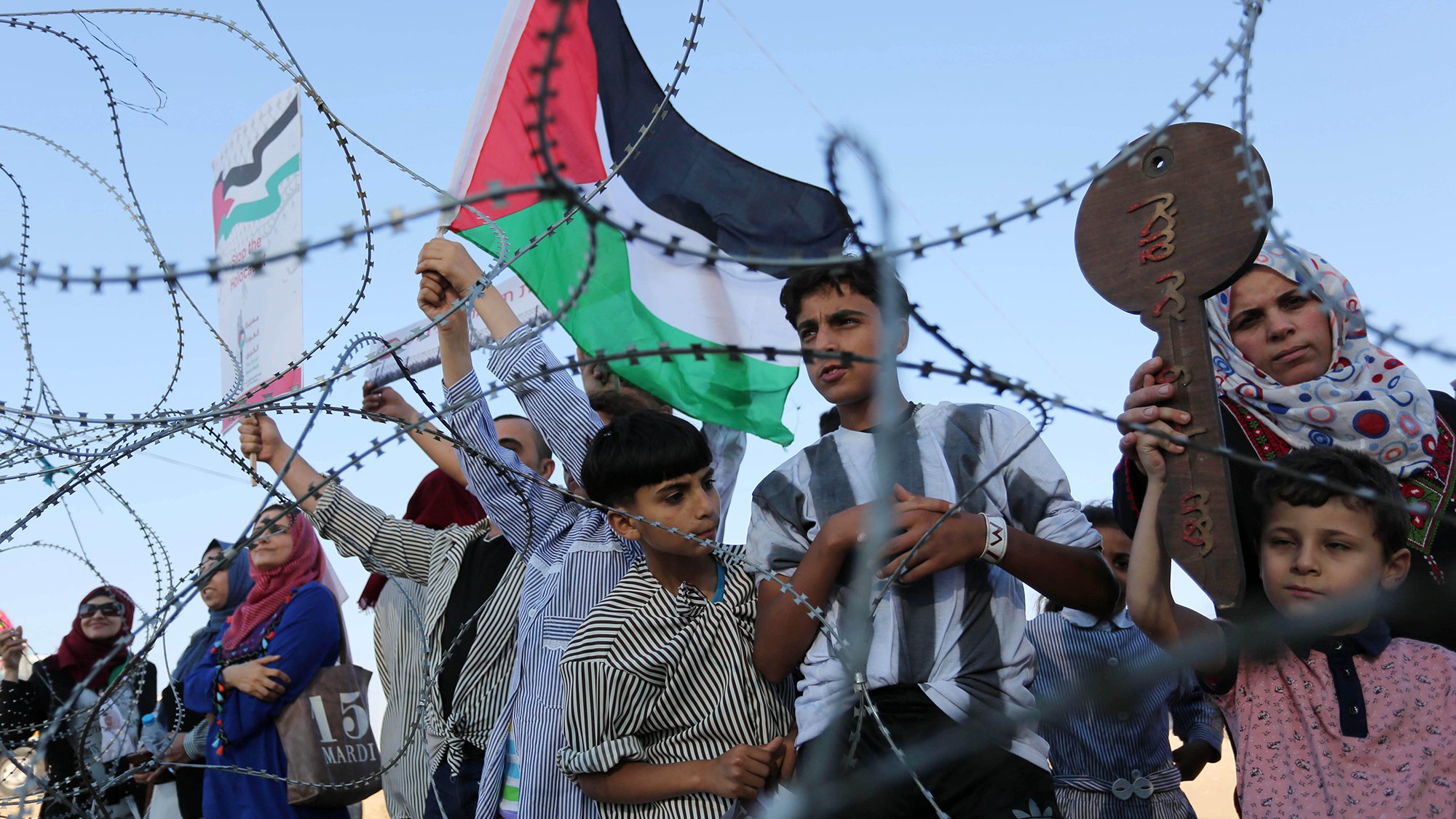 در این تصویر، گروهی از مردم، از جمله کودکانی، در نزدیکی یک نوار سیم‌خاردار تجمع کرده‌اند. برخی از آن‌ها پرچم فلسطین را در دست دارند و به نظر می‌رسد که در حال ابراز احساسات یا مخالفت با شرایطی هستند. لباس‌های مختلفی بر تن دارند و حالت‌هایشان متنوع است. زمینه تصویر آسمانی صاف و روشن به نظر می‌رسد.