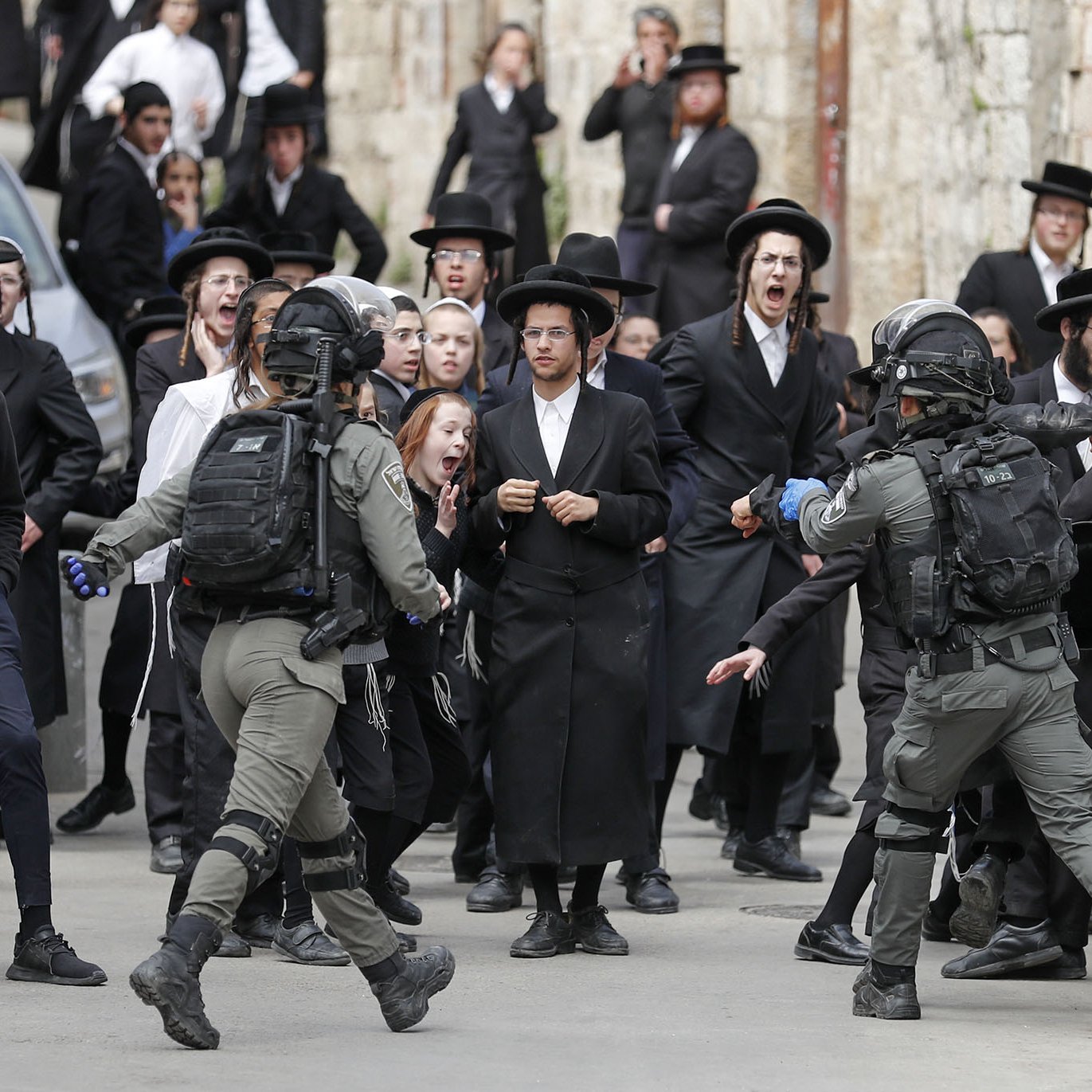 L'image montre une scène animée dans une rue, où un groupe de personnes, principalement habillées en noir, semble manifester ou réagir à une situation tendue. On peut voir des hommes en habits traditionnels, portant des chapeaux noirs, ainsi que des adolescents sur des bicycles. Au centre, des membres des forces de l'ordre, en tenue tactique, interagissent avec la foule. Les expressions faciales des personnes dans la foule varient entre la surprise, l'inquiétude et l'agitation, créant une atmosphère de tensions. Les bâtiments en arrière-plan évoquent un contexte urbain, probablement dans une région où se concentrent des communautés religieuses.