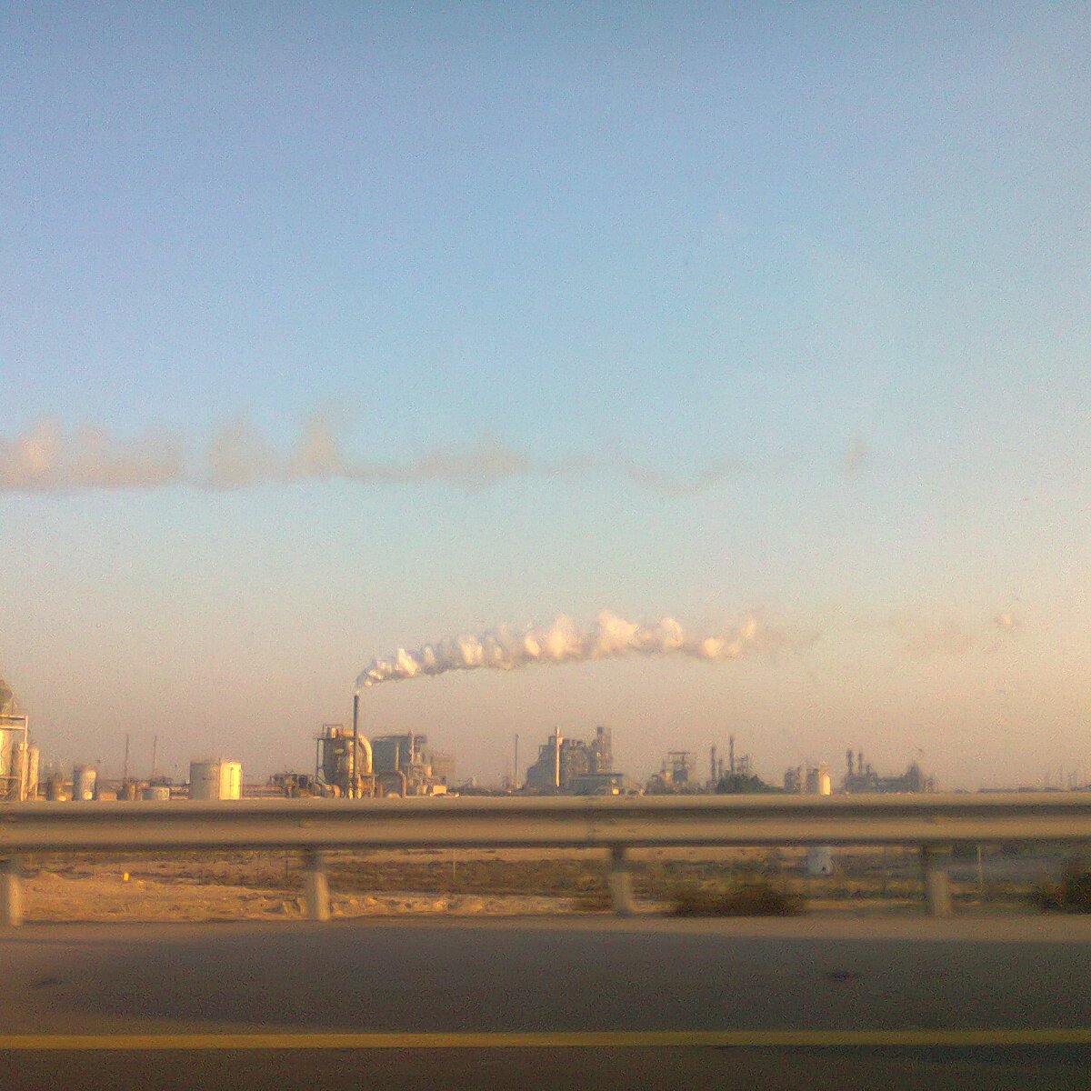 L'image montre un paysage industriel avec plusieurs structures d'usine. On voit de grandes cheminées qui émettent des nuages de fumée blanche dans le ciel. Le ciel est clair, avec une lumière douce, ce qui donne une atmosphère tranquille malgré l'activité industrielles. Au premier plan, on aperçoit une route avec une barrière en béton, indiquant que l'on observe ce paysage en passant en véhicule. L'ensemble dégage une impression de production industrielle dans un environnement ouvert.