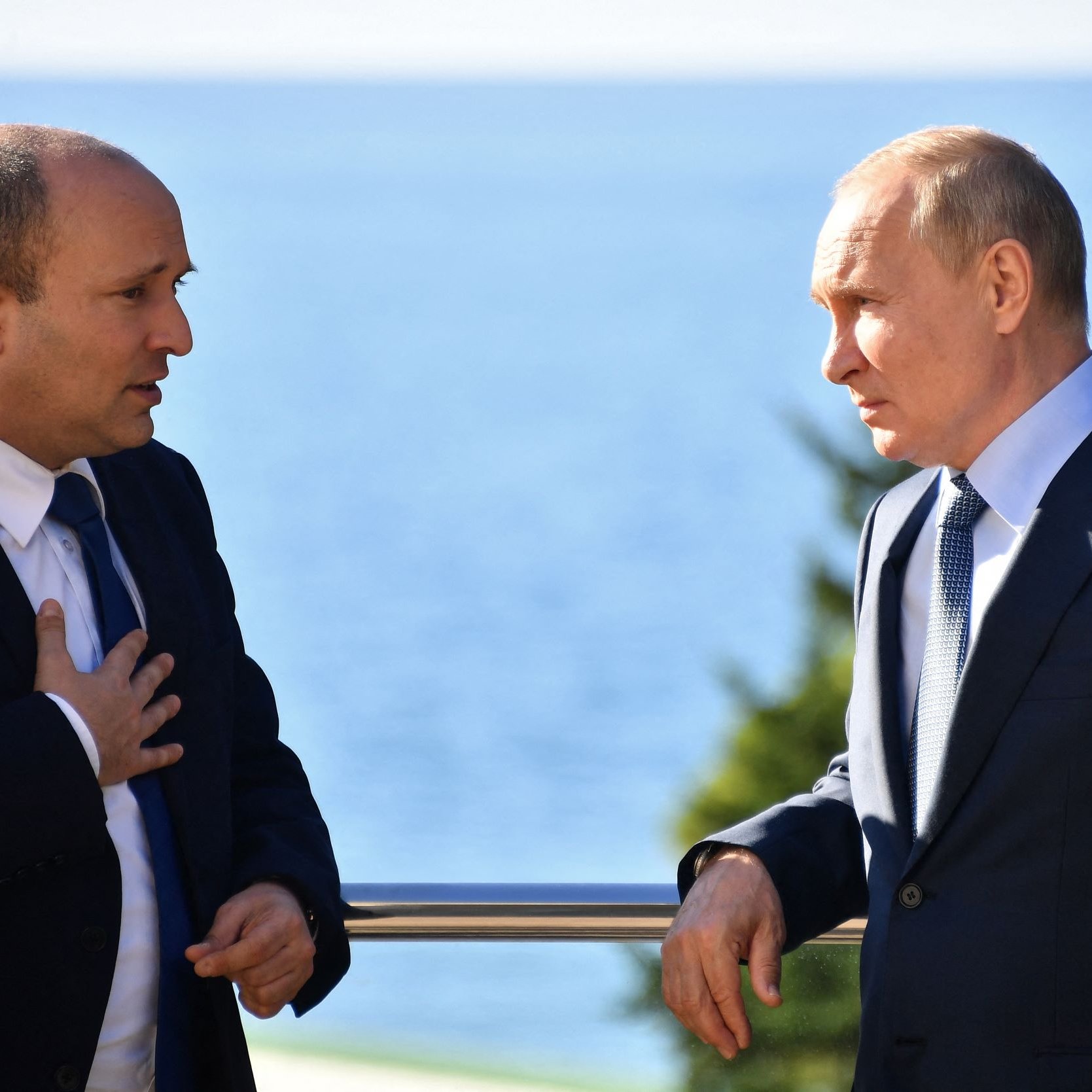 L'image montre deux hommes en discussion près d'une balustrade, avec une vue sur la mer en arrière-plan. L'un d'eux, portant une kippa, semble s'exprimer avec gestuelle, tandis que l'autre écoute attentivement. Les deux hommes sont habillés de manière formelle, et l'atmosphère paraît concentrée. La lumière du jour éclaire la scène, suggérant un cadre extérieur agréable.