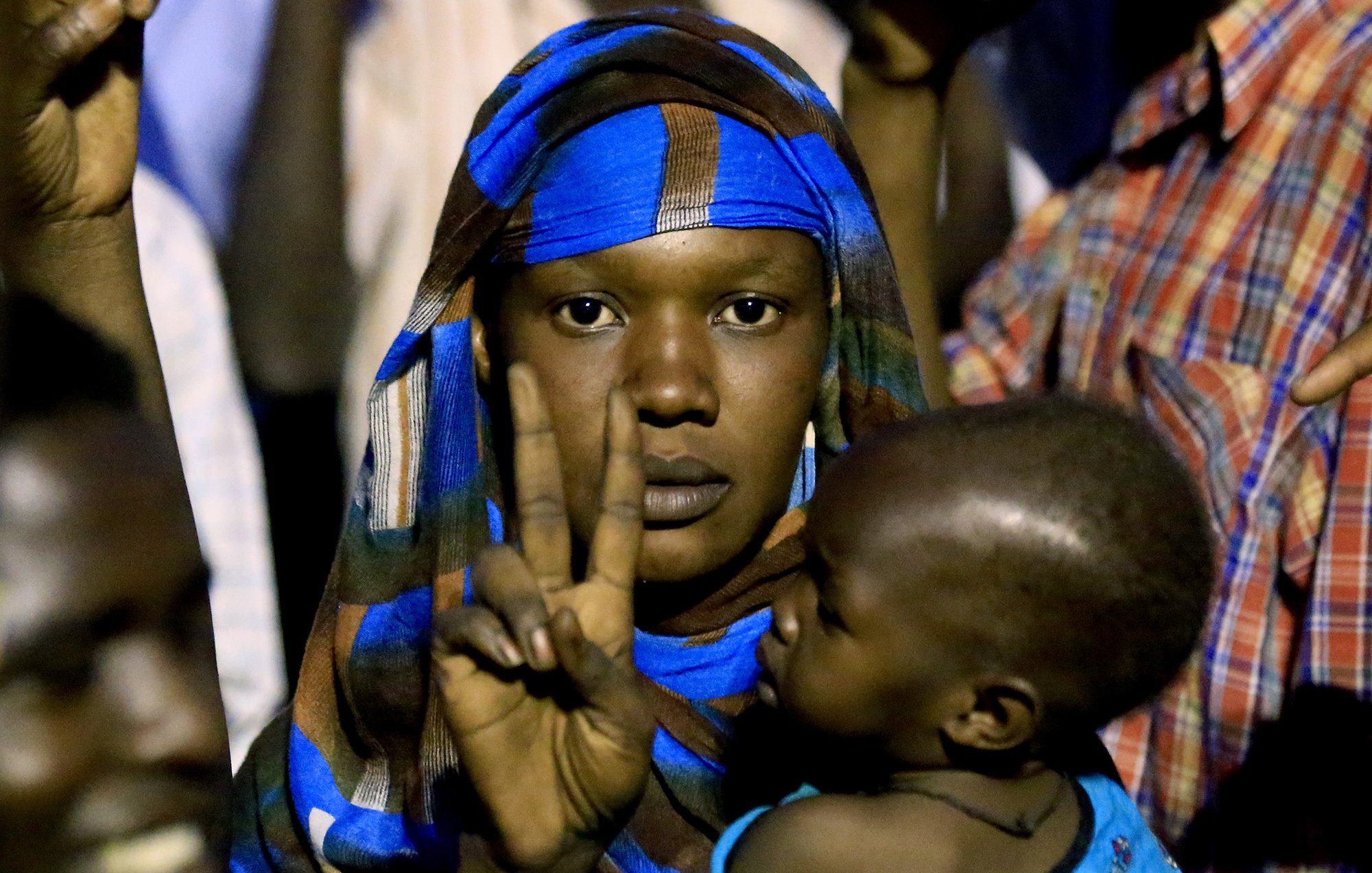L'image montre une femme portant un foulard coloré, qui fait un signe de paix avec sa main. Elle tient un jeune enfant dans ses bras. L'arrière-plan semble flou, suggérant qu'elle se trouve dans une foule. Les expressions sur son visage et sa posture peuvent indiquer un moment de détermination ou de solidarité.