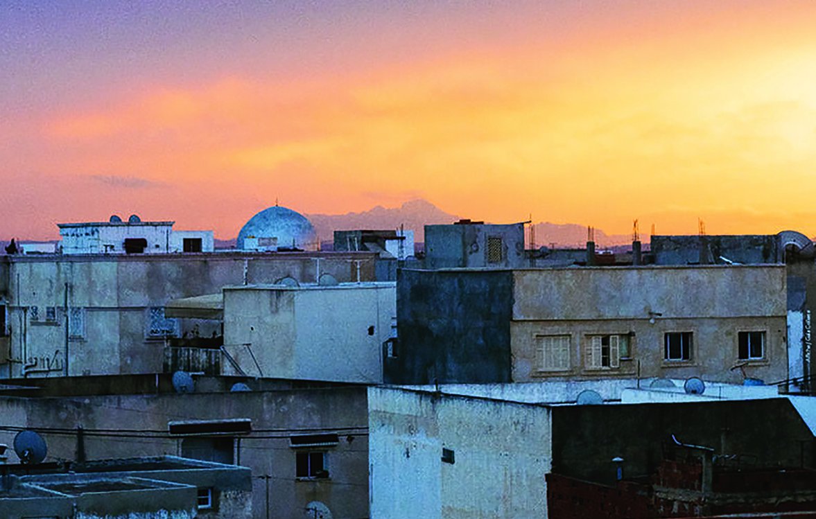 L'image montre un paysage urbain au coucher du soleil. On y voit plusieurs bâtiments aux façades de couleur beige et grise, avec des toits plats. À l'horizon, une belle teinte orange et rose illumine le ciel, créant une ambiance chaleureuse. Un dôme se distingue parmi les structures, suggérant la présence d'une mosquée ou d'un autre bâtiment significatif. Les lignes des constructions et le ciel coloré contrastent magnifiquement, donnant une impression de tranquillité.