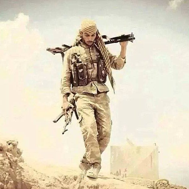 L'image montre un homme portant une tenue militaire, avec un chèche sur la tête. Il marche sur un terrain désertique, tenant deux armes, l'une à la main et l'autre portée sur son épaule. Son expression semble déterminée et il est entouré d'un paysage aride. L'atmosphère de l'image évoque un contexte de conflit ou de guerre.