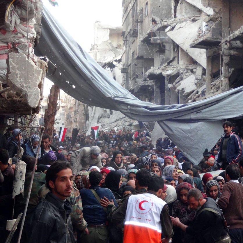 L'image montre une scène de destruction massive dans une zone urbaine, probablement après un conflit ou une catastrophe. On peut voir des bâtiments gravement endommagés, avec des murs effondrés et des débris partout. Une foule de personnes, dont certaines semblent déplacées, se rassemble dans la rue. Il y a des tentes et des bâches qui servent d'abris temporaires. Des membres d'organisations humanitaires, identifiables par leurs vêtements distinctifs, sont présents pour aider. L'ambiance générale est celle de la détresse et de la recherche d'aide dans une situation de crise.