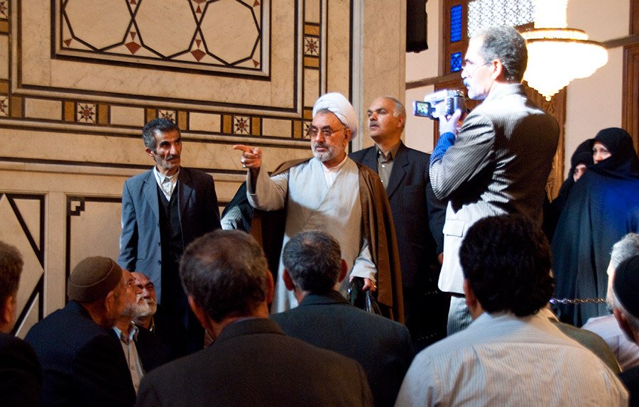 L'image montre un groupe de personnes rassemblées dans un espace intérieur richement décoré. Au centre, un homme en vêtements traditionnels, probablement religieux, semble s'adresser à l'assemblée. Il lève une main en signe d'argumentation ou d'expression. Plusieurs autres hommes l'écoutent attentivement, tandis qu'une ou deux femmes en arrière-plan portent un hijab. L'atmosphère semble sérieuse et respectueuse, avec des détails architecturaux élégants autour.