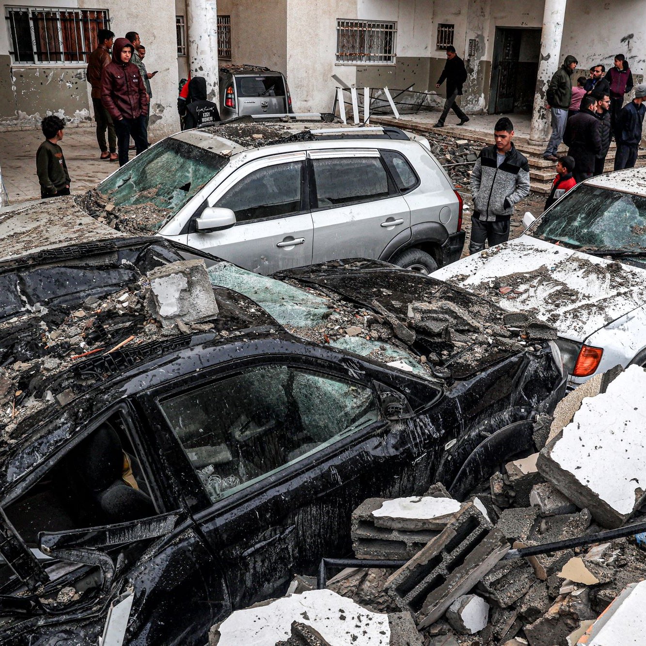 این تصویر نشان‌دهنده‌ یک صحنه ویران شده به واسطه زلزله است. چندین خودرو زیر آوارهای ساختمانی قرار گرفته‌اند و آسیب زیادی دیده‌اند. افرادی در پس‌زمینه در حال مشاهده و بررسی وضعیت هستند. آوار خانه‌ها و دیوارهای فروریخته در اطراف خودروها به چشم می‌خورد. به طور کلی، جو تصویر نشان‌دهنده اضطراب و فاجعه ناشی از زلزله است.