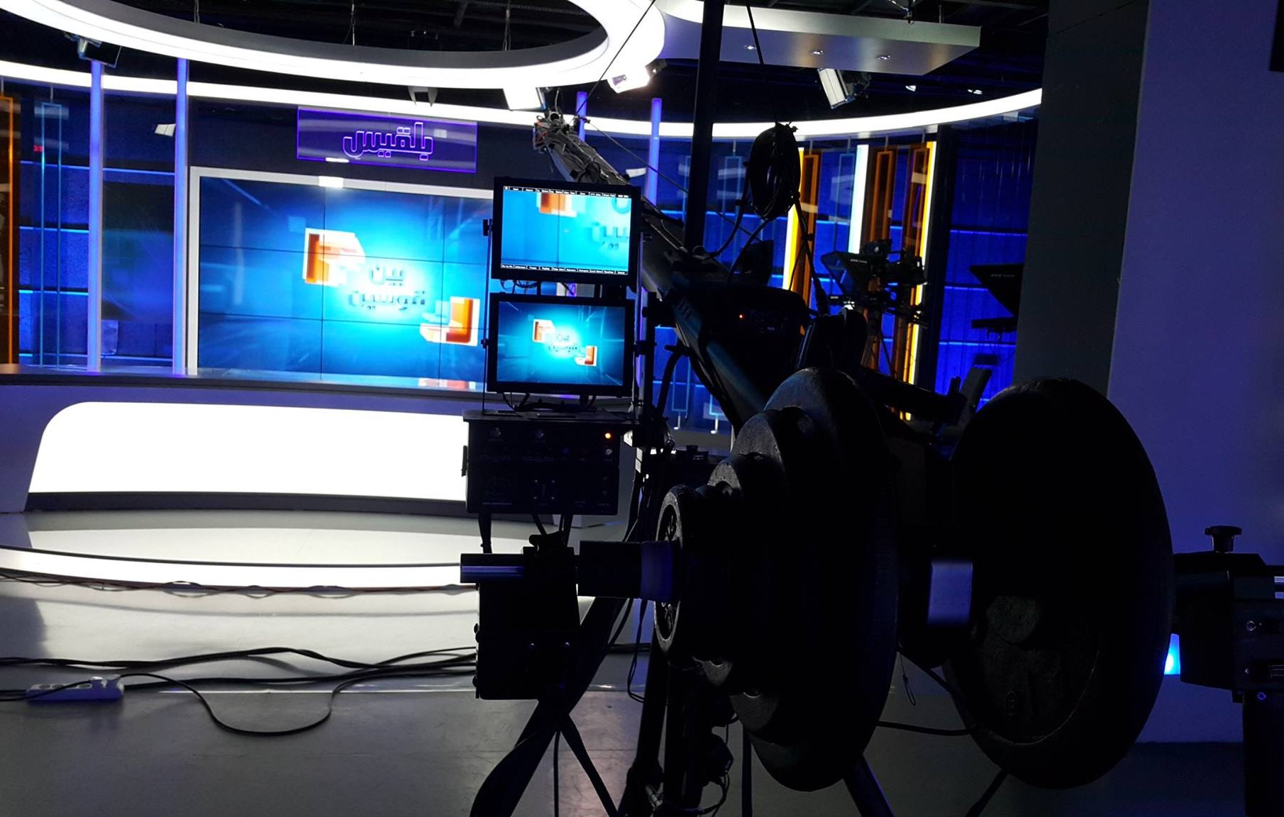 L'image montre un studio de télévision moderne. On peut y voir des lumières bleues qui éclairent l'espace, avec un fond décoratif et des écrans de présentation. À l'avant-plan, il y a une caméra sur un trépied, ainsi que des équipements techniques, ce qui indique que le studio est préparé pour une émission en direct ou un enregistrement. L'ambiance semble professionnelle et bien équipée pour la diffusion.