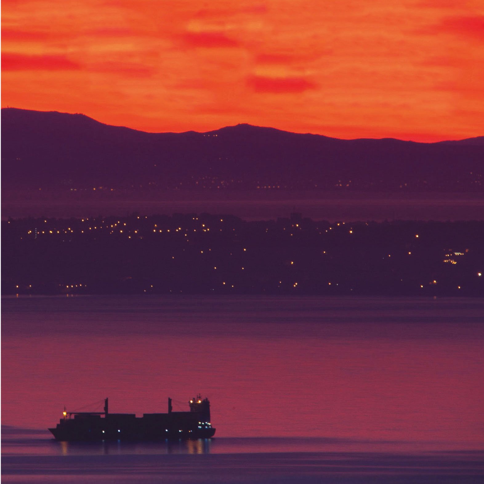 L'image présente un coucher de soleil spectaculaire, où le ciel est teinté de rouge et d'orange lumineux. En arrière-plan, on aperçoit des collines sombres, tandis qu'à l'avant-plan, un cargo navigue sur l'eau calme. Les lumières d'une ville ou d'un port scintillent le long de la côte, ajoutant une touche de vie à cette scène paisible.