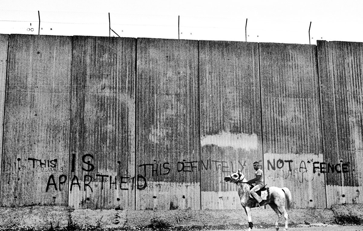 L'image montre un cavalier monté sur un cheval, se tenant près d'un grand mur de béton. Ce mur est marqué par des graffitis en anglais, indiquant des messages provocateurs tels que "THIS IS APARTHEID" et "THIS DEFINITELY NOT A FENCE". L'ambiance de la photo souligne un contraste entre la scène rurale du cavalier et la nature austère du mur, évoquant des thèmes de séparation et de conflit.