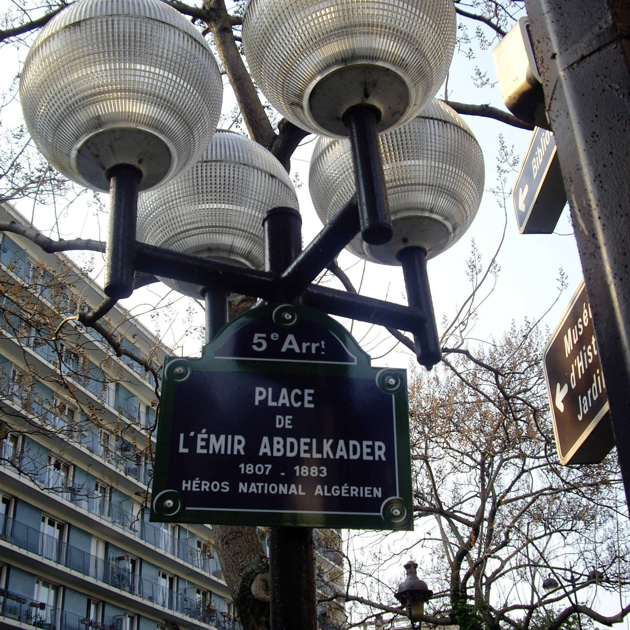 L'image montre un lampadaire avec plusieurs globes lumineux, sur lequel est fixée une plaque signalétique. Cette plaque indique "Place de l'Émir Abdelkader", accompagnée des dates 1807-1883 et de la mention "Héros National Algérien". Le tout se situe dans le cinquième arrondissement, comme le précise le petit panneau en haut. En arrière-plan, on peut apercevoir des bâtiments typiques de la ville.
