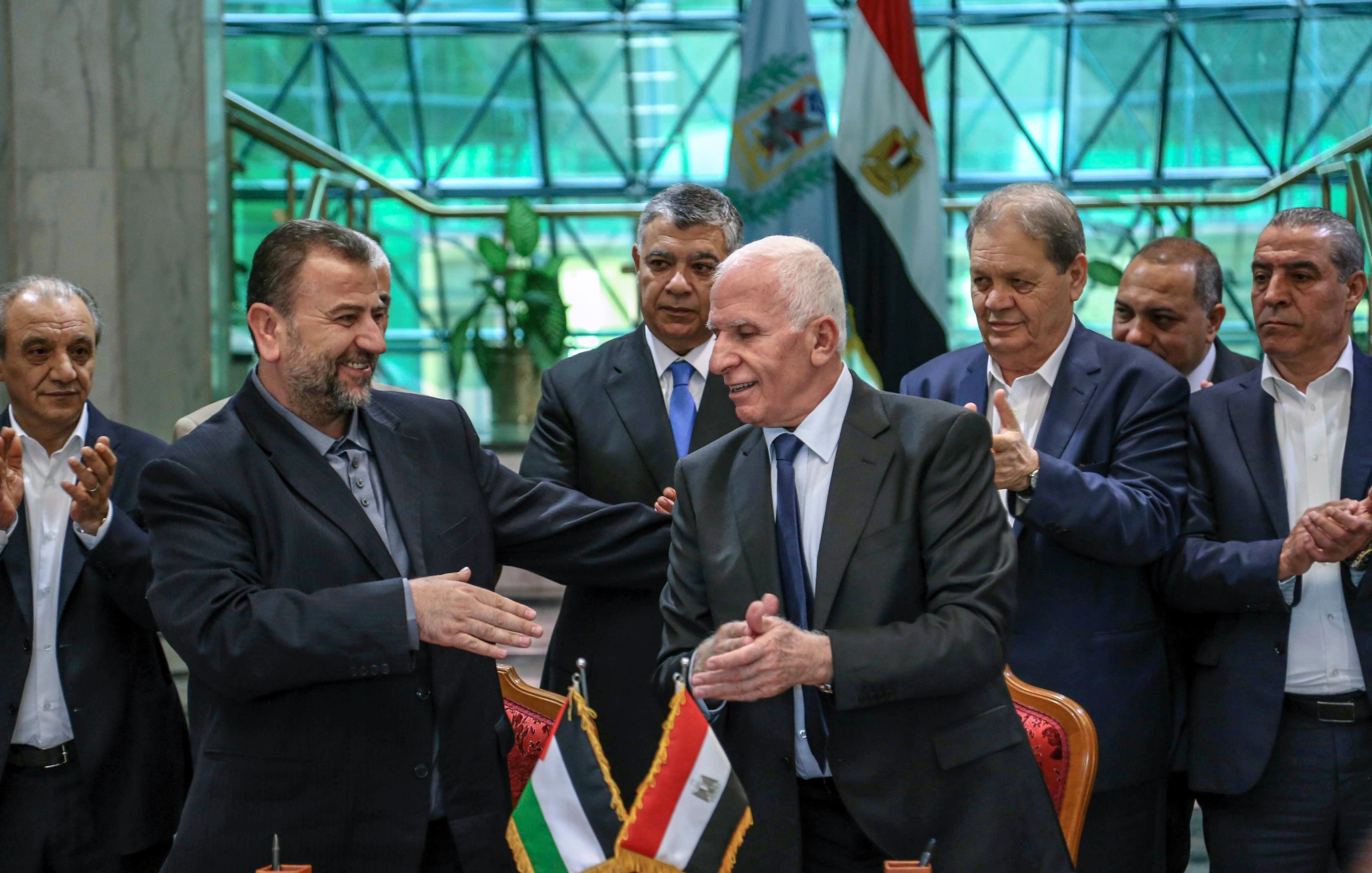 The image shows a formal gathering where two men are shaking hands, likely after a significant agreement or event. They are standing at a table with flags of Palestine and Egypt displayed in front of them. Behind them, a group of men appears to be applauding or showing support. The setting looks official, possibly within a government building, with a modern architectural backdrop. The atmosphere seems positive, indicating a moment of cooperation or partnership.
