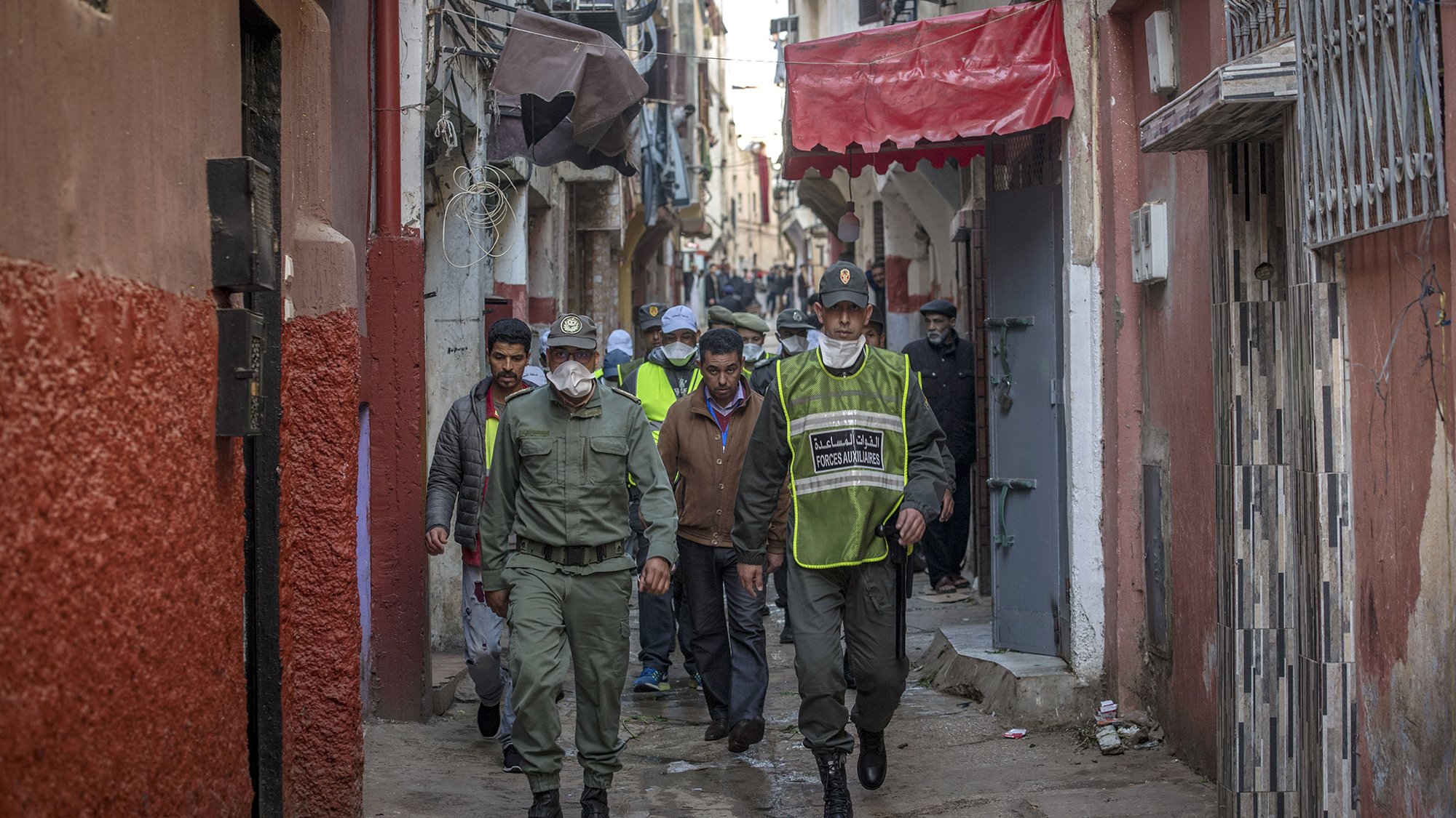 L'image montre une ruelle étroite et animée, typique d'une ville marocaine. Au premier plan, des agents de police ou des membres de la sécurité patrouillent. Ils portent des uniformes et des masques, indiquant une attention à la sécurité ou à la santé publique. En arrière-plan, on peut voir des habitants et d'autres personnes dans la rue, ainsi que des bâtiments aux façades colorées. L'atmosphère semble à la fois dynamique et surveillée, reflétant la vie quotidienne dans ce type d'environnement urbain.