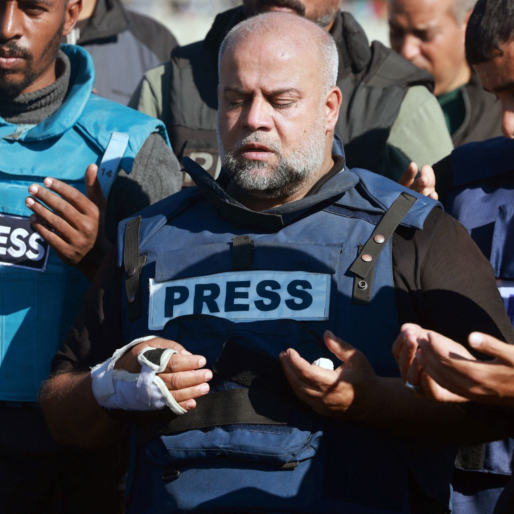 L'image montre un groupe de personnes, dont un homme au centre, portant un gilet avec l'inscription "PRESS". Cet homme semble prier ou méditer, ayant les yeux fermés. Il a également un bandage sur une main. Les autres personnes autour semblent également impliquer dans un moment de recueillement. Le contexte semble émotionnel ou solennel, suggérant une situation liée à des événements récents.