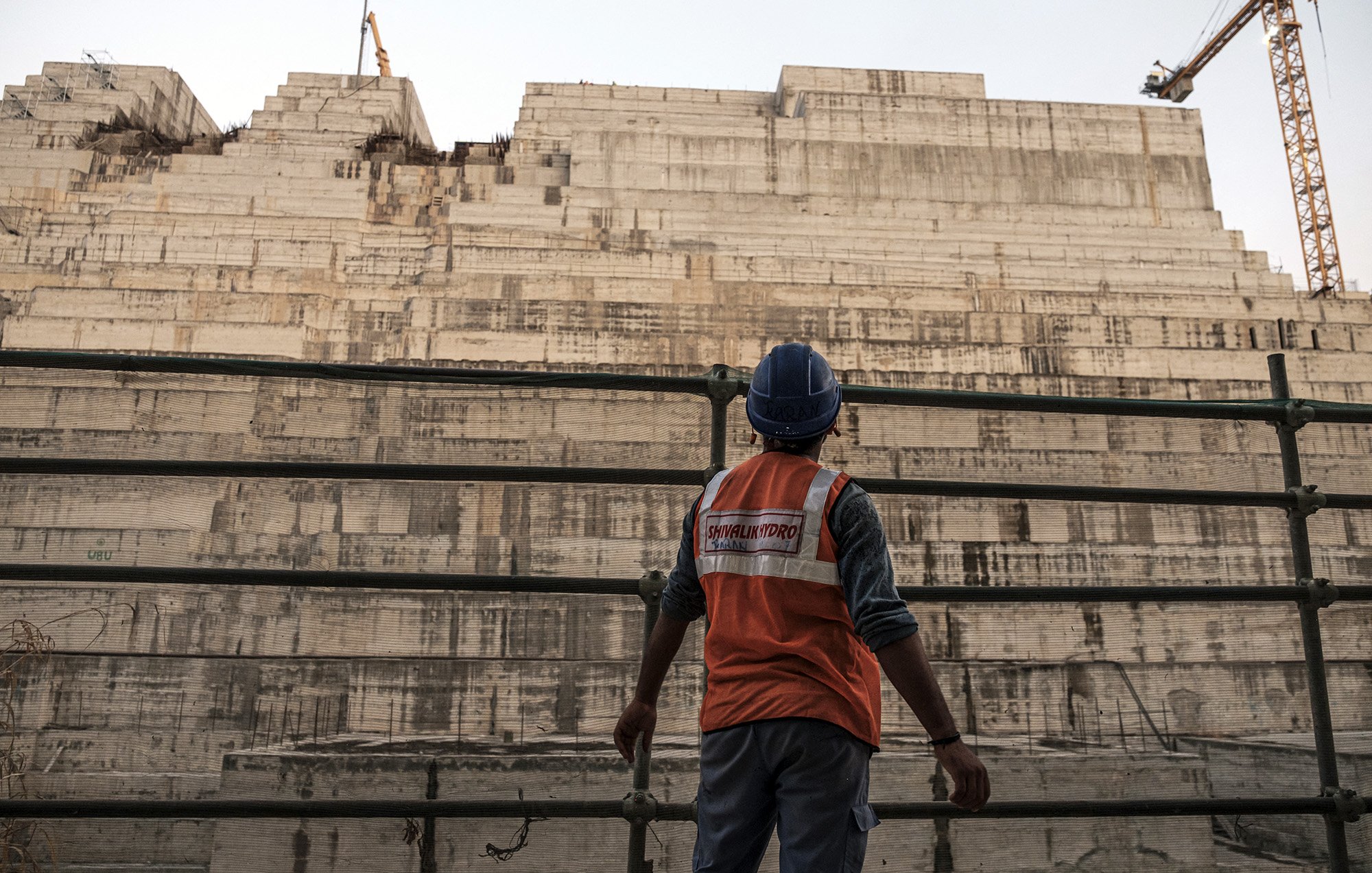 L'image montre un ouvrier sur un chantier de construction, portant un gilet de sécurité orange et un casque. Il se tient devant une grande structure en béton, apparemment inachevée, qui est composée de plusieurs niveaux superposés. En arrière-plan, on aperçoit une grue, ce qui indique que le site est actif. L'atmosphère semble à la fois industrielle et impressionnante, mettant en avant l'ampleur du projet de construction.