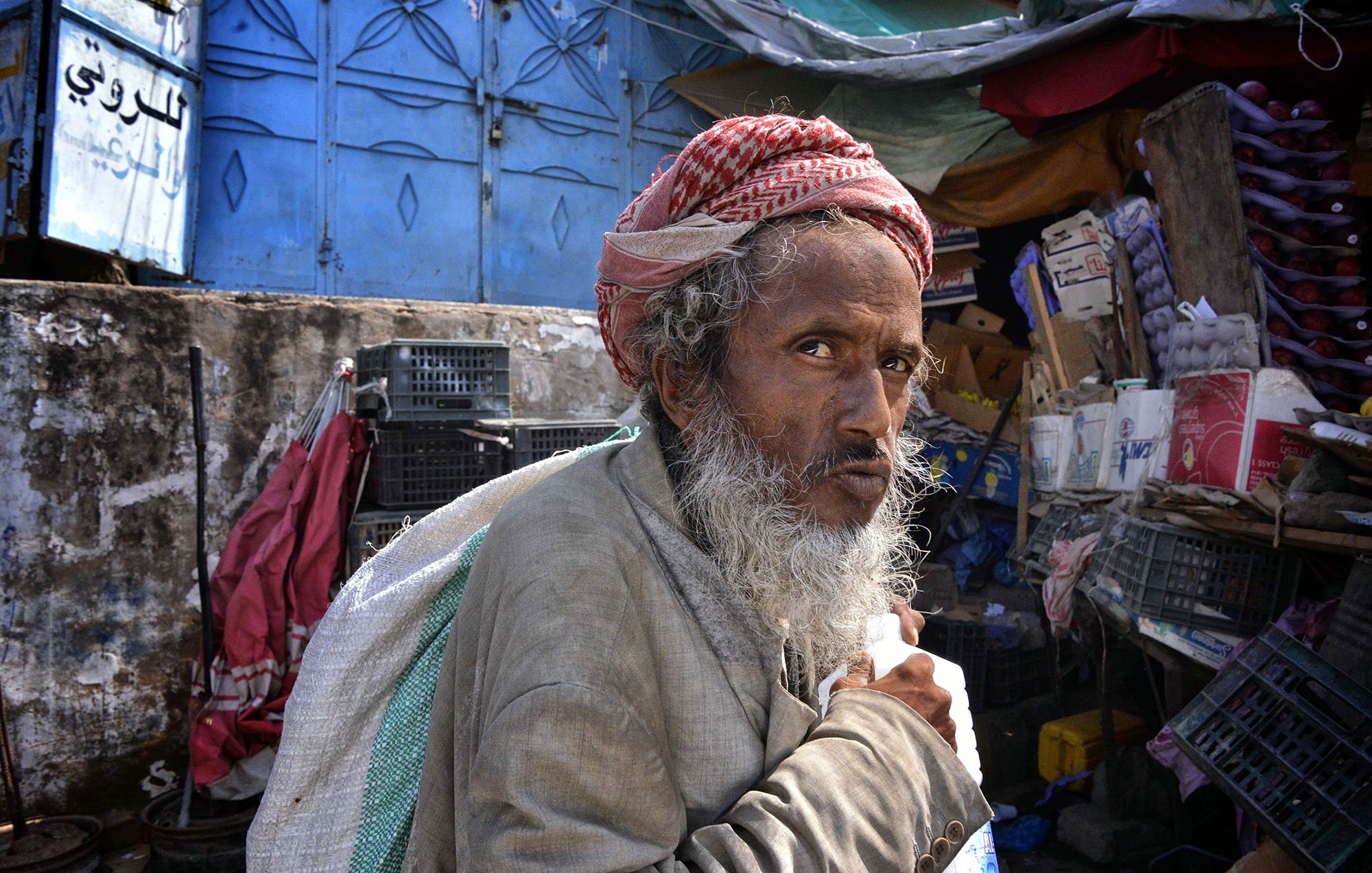 L'image montre un homme âgé avec une longue barbe grise, portant un turban rouge et blanc. Il semble regarder fixement avec une expression pensante ou préoccupée. L'arrière-plan suggère un marché ou un bazar, avec des étals et des objets divers. L'éclairage est naturel, ce qui donne une atmosphère vivante à la scène. L'homme porte un sac, ce qui indique qu'il pourrait être en train de se déplacer ou de faire des courses.