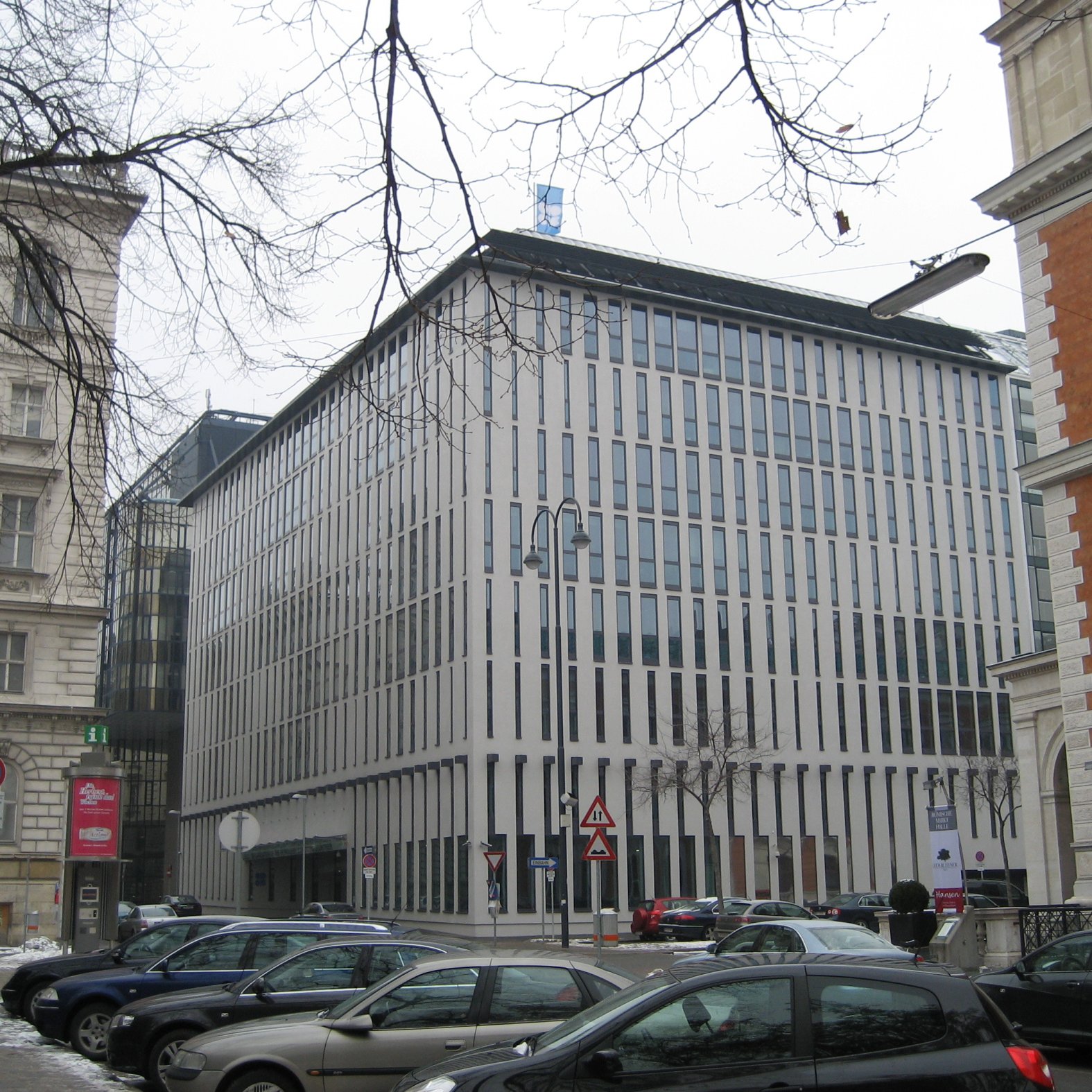 این تصویر نمای یک ساختمان مدرن را نشان می‌دهد که دارای طراحی مینیمالیستی است. نمای ساختمان با پنجره‌های عمودی و خطوط باریک تقسیم شده، ظاهری مرتب و شیک دارد. اطراف ساختمان ممکن است شامل خودروها و درختان بدون برگ در فصل زمستان باشد. در سمت چپ و راست تصویر، ساختمان‌های دیگری با طراحی‌های کلاسیک‌تر وجود دارند که تضادی جالب با ساختمان مدرن ایجاد می‌کنند. فضا به نظر می‌رسد که در یک منطقه شهری و ممکن است در فصل سرد سال باشد.