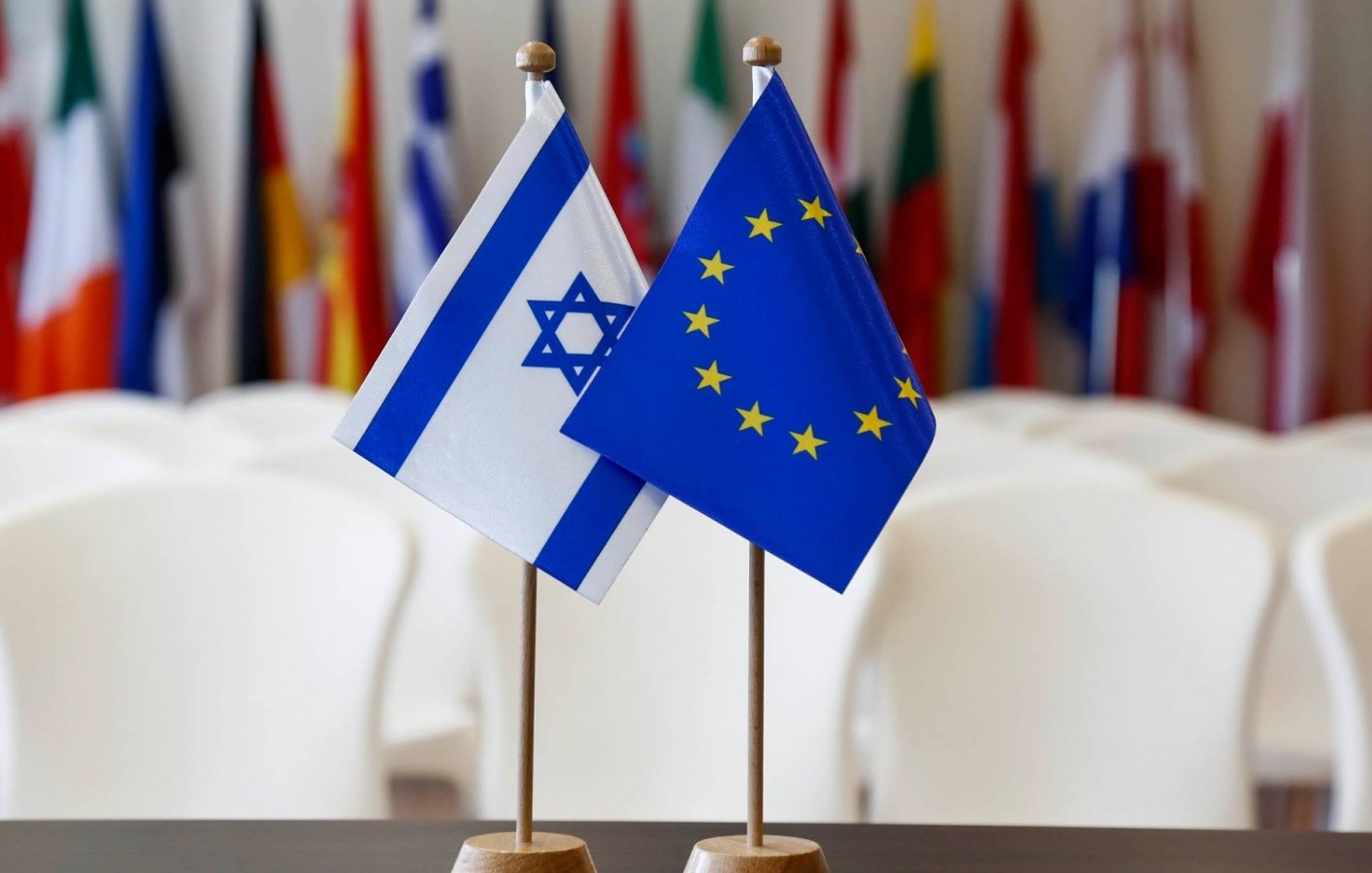 L'image montre deux drapeaux placés sur des supports. À gauche se trouve le drapeau d'Israël, qui présente une étoile de David et deux bandes bleues. À droite se trouve le drapeau de l'Union européenne, emblématique avec des étoiles dorées disposées en cercle sur un fond bleu. En arrière-plan, on aperçoit un rassemblement de drapeaux de différents pays, suggérant un contexte international ou diplomatique. Les chaises blanches sont disposées derrière, évoquant une salle de réunion ou une conférence.