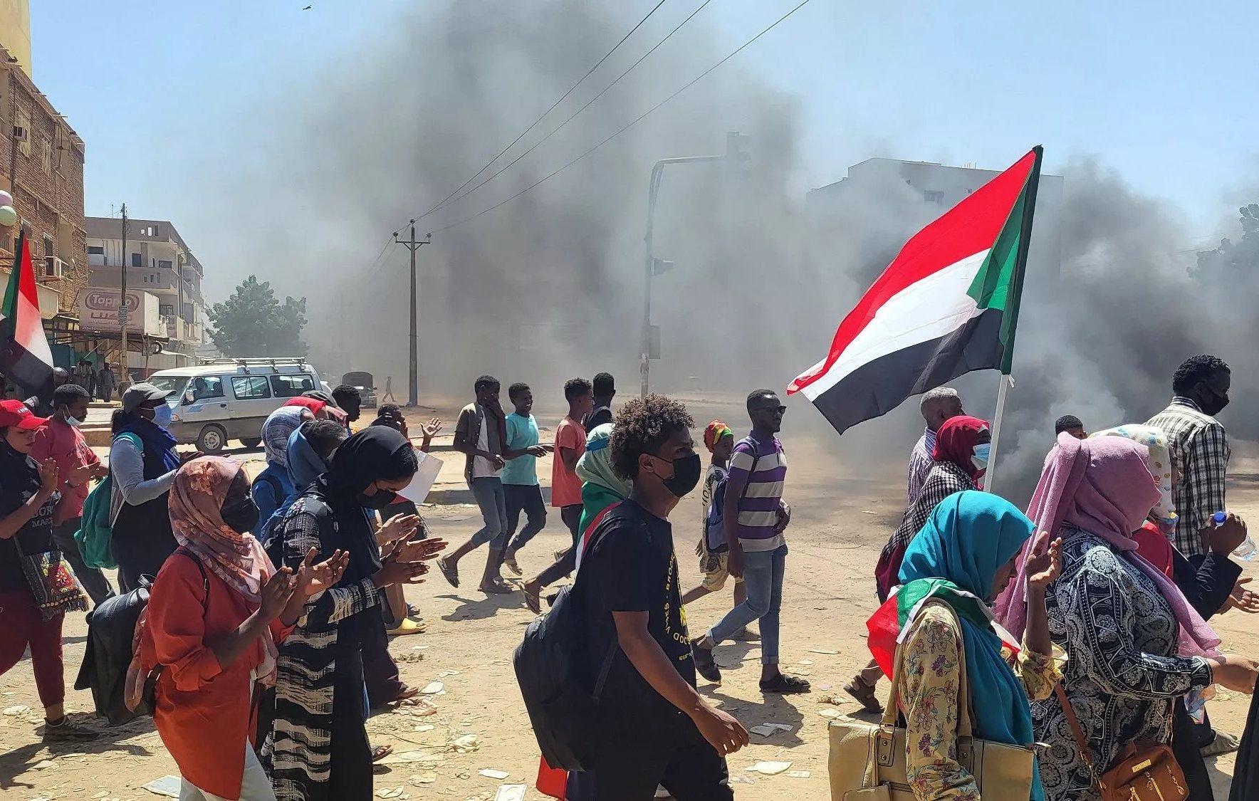 Manifestanti con bandiera del Sudan, in mezzo a fumi di incendi e tensione.