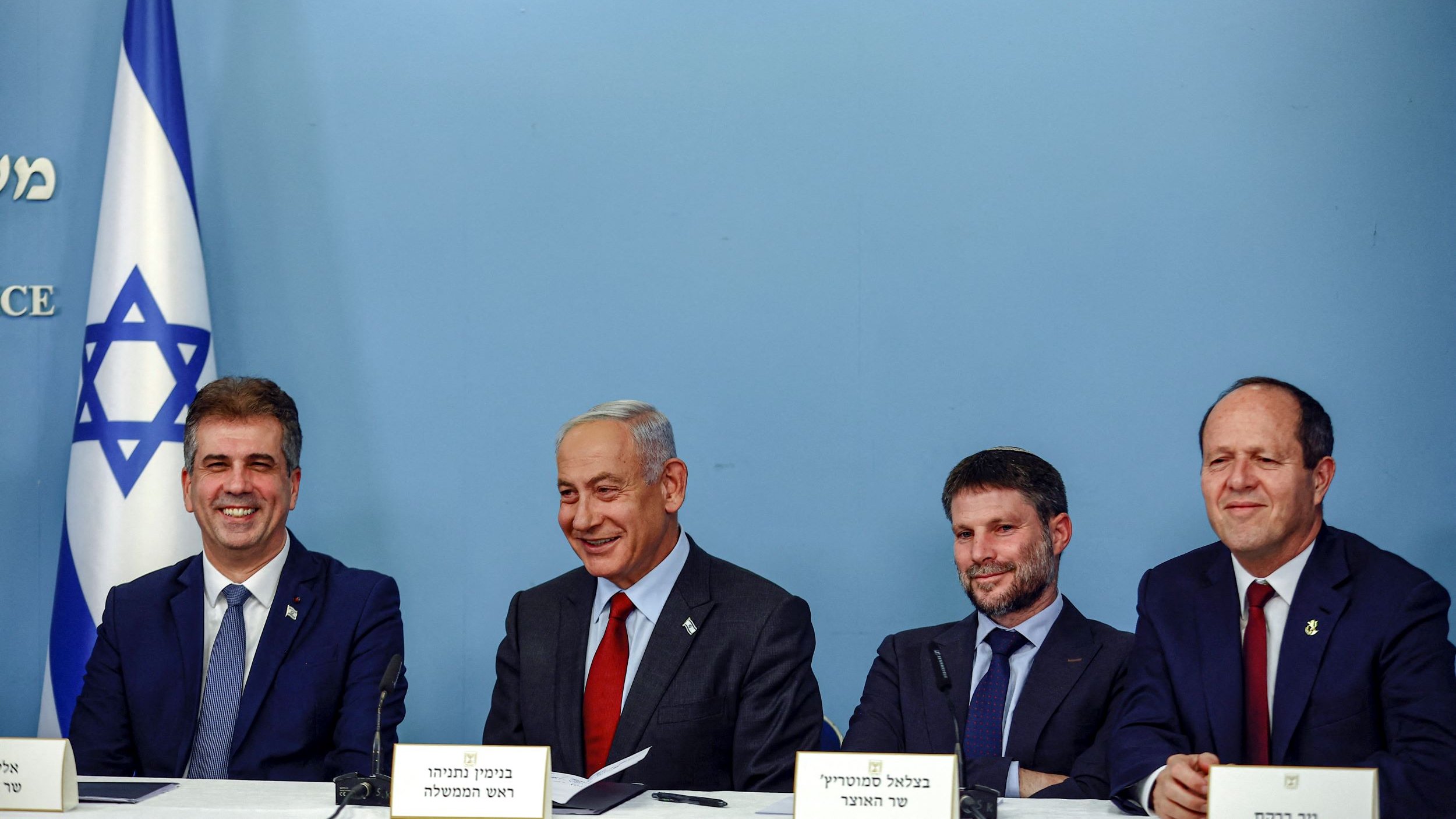 L'image montre un groupe de quatre hommes assis à une table, souriant et discutant. En arrière-plan, il y a un drapeau israélien. Les hommes portent des costumes et des cravates, et il semble qu'ils soient en train de participer à une conférence de presse ou à un événement officiel. L'ambiance est décontractée mais professionnelle.