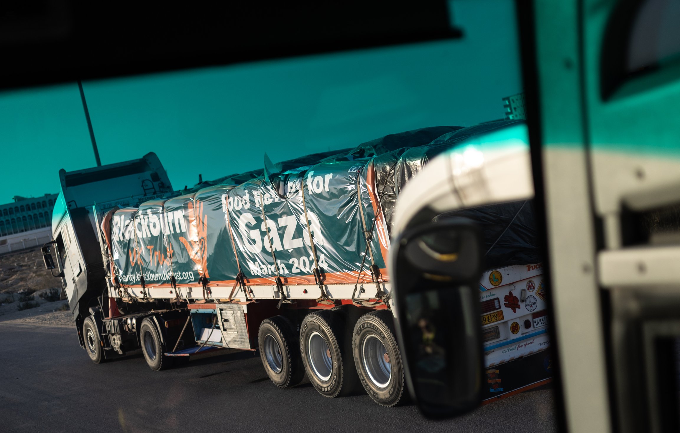 L'immagine mostra un camion lungo che trasporta una grande quantità di beni. Il veicolo è visibile da un angolo diverso, suggerendo che la foto potrebbe essere stata scattata da un altro veicolo. Sulla superficie del camion è presente un'informazione, probabilmente un messaggio di solidarietà o supporto, che menziona "Gaza" e "marzo 2024". Il contesto suggerisce un'operazione umanitaria o di aiuto. Sullo sfondo si possono vedere elementi di un paesaggio stradale.