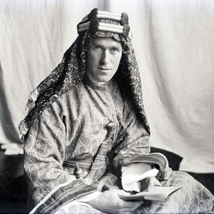 L'image montre un homme portant un costume traditionnel. Il est vêtu d'une tunique ornée et d'un headdress décoratif. Son regard est sérieux et il tient un livre dans ses mains. L'arrière-plan est sobre, ce qui met en valeur son apparence et son habillement. La photo semble être ancienne, ce qui lui donne un aspect historique.