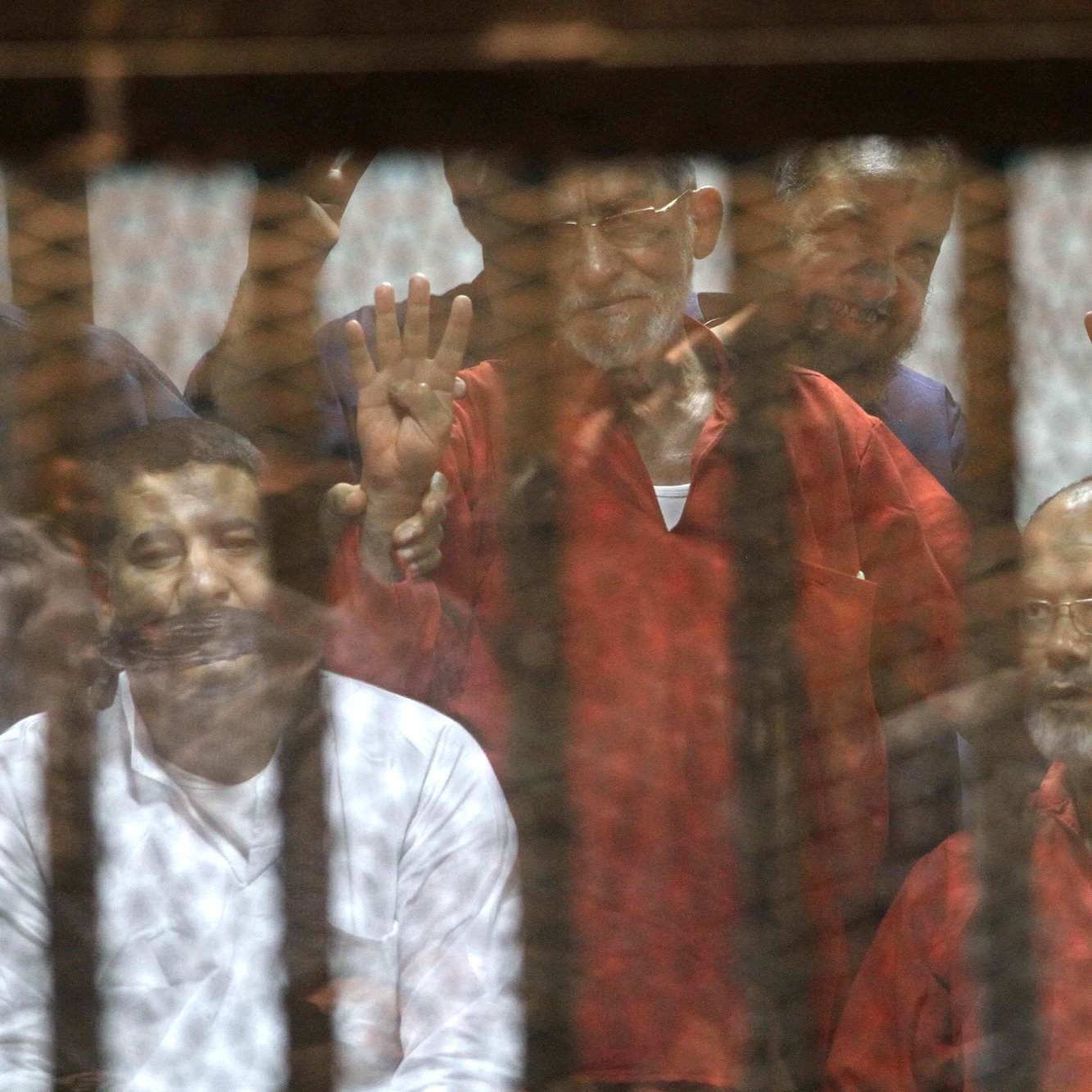 تظهر في الصورة مجموعة من الأشخاص الموجودين خلف قضبان. يبدو أن بعضهم يرفعون إشارة معينة، بينما تعكس الشبكة المحيطة بهم بعض التفاصيل، مما يعطي انطباعًا عن مجموعة محكومة في بيئة موصولة. الألوان تتراوح بين الأحمر والأبيض، مما يشير إلى ملابس يمكن أن تكون متعلقة بسياق خاص. الوضع العام يوحي بالتوتر أو الأحداث الجادة.