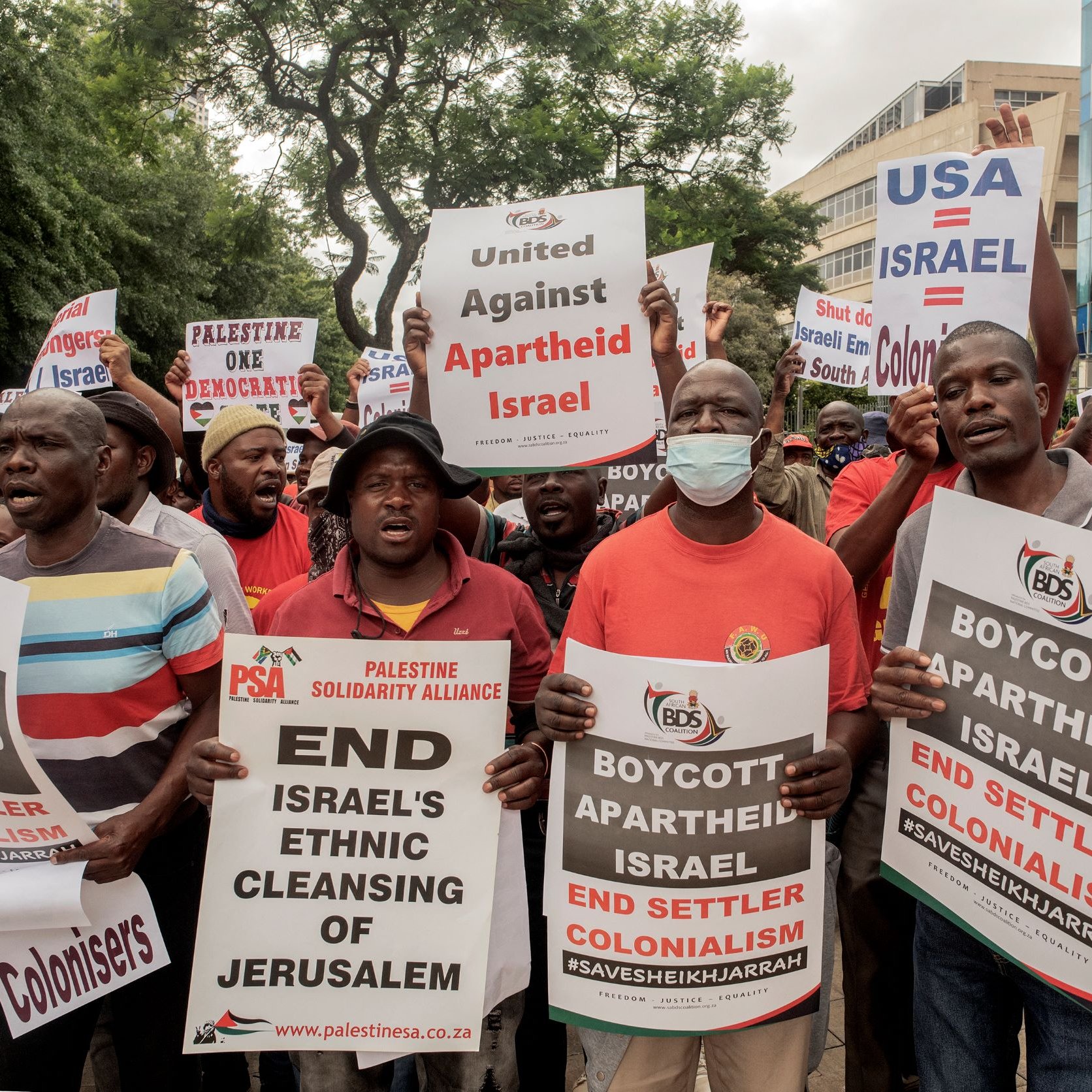 L'immagine ritrae un gruppo di manifestanti impegnati in una protesta. I partecipanti, vestiti in gran parte con magliette rosse, tengono in mano cartelli con testi che chiedono la fine dell'apartheid in Israele, il boicottaggio delle politiche israeliane e l'unità contro il colonialismo. Alcuni cartelli menzionano specificamente il "Pulizia etnica di Gerusalemme" e fanno riferimento ad azioni di protesta contro l'ambasciata degli Stati Uniti in relazione a questi temi. L'atmosfera sembra carica di energia e determinazione.