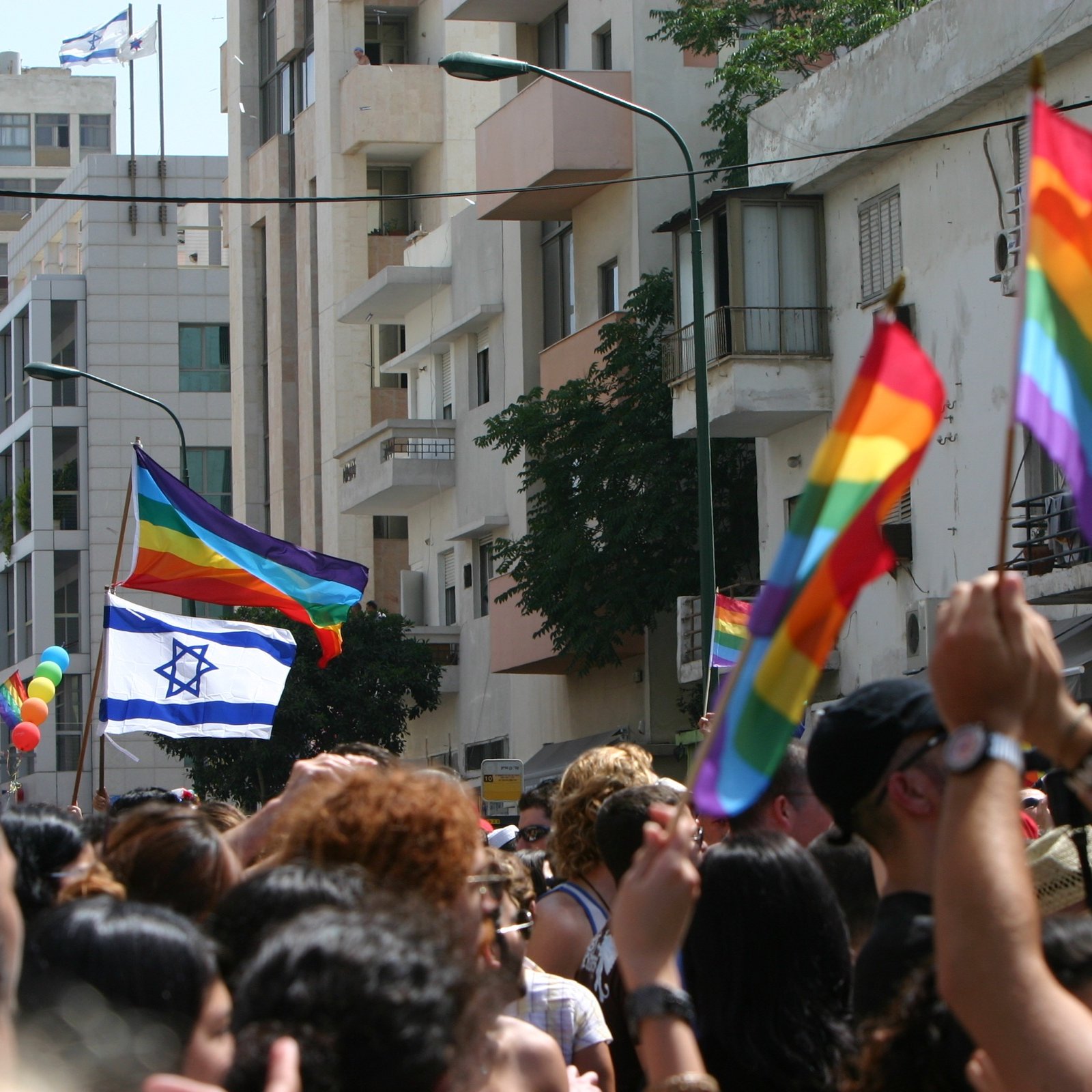 L'image montre une scène vibrante de célébration lors d'un défilé de fierté. On y voit une foule de personnes brandissant des drapeaux arc-en-ciel, symboles de la communauté LGBTQ+. Parmi eux, une grande bannière représentant le drapeau d'Israël flotte également. En arrière-plan, on distingue des bâtiments urbains, et plusieurs personnes semblent s'amuser et célébrer l'événement, créant une ambiance festive et joyeuse.