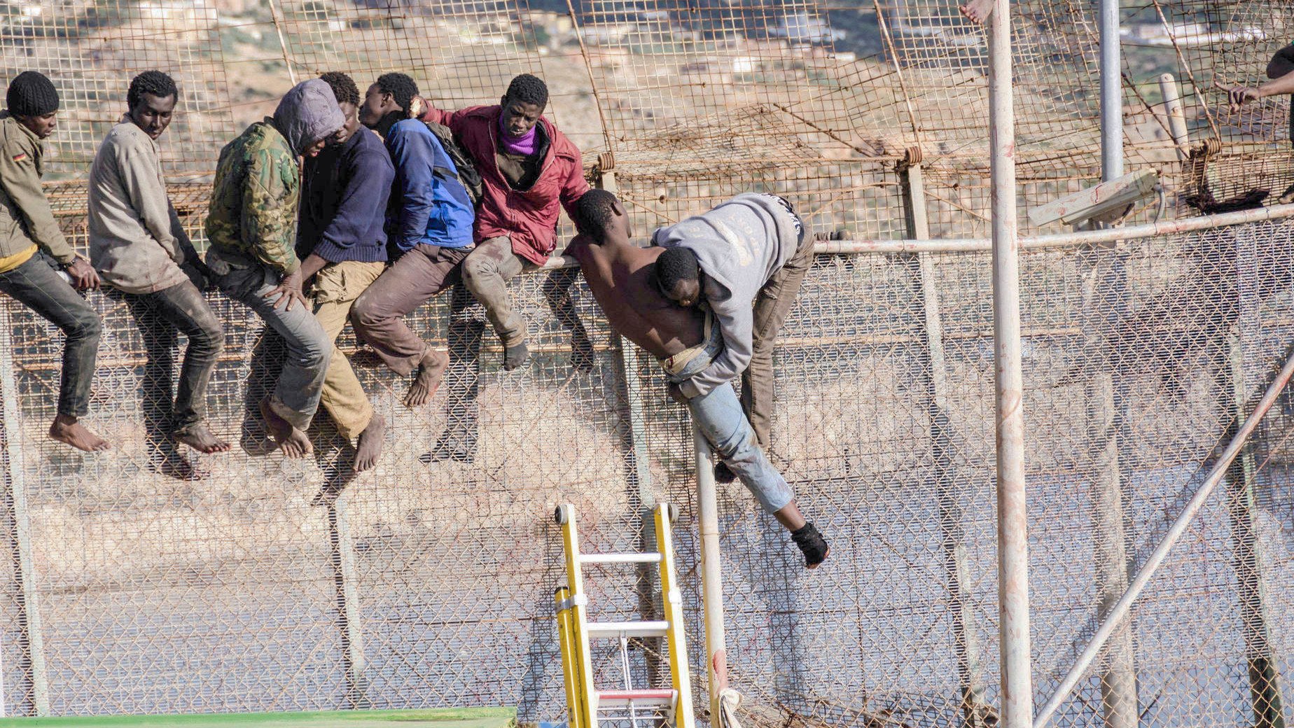 L'image représente un groupe de personnes tentant de franchir une clôture métallique. On peut voir plusieurs individus en train de grimper et de passer par-dessus la barrière, certains sont déjà en train de descendre de l'autre côté. L'environnement semble être une zone frontalière, probablement dans un contexte de migration. Les personnes semblent déterminées, et l'image transmet une forte impression d'urgence et de mouvement.