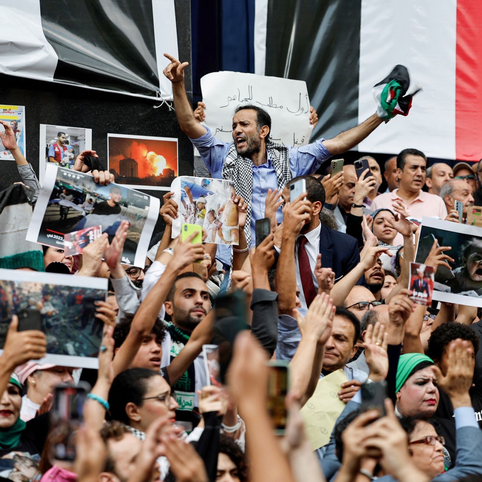 L'image montre une foule rassemblée lors d'une manifestation, avec des personnes brandissant des pancartes et des photos. Au centre, un homme lève les bras en signe de victoire ou de protestation. La foule exprime visiblement des émotions fortes, que ce soit la colère ou la solidarité. Des drapeaux sont également présents dans le décor, ajoutant à l'intensité de la scène. On ressent une atmosphère d'engagement et de mobilisation collective.