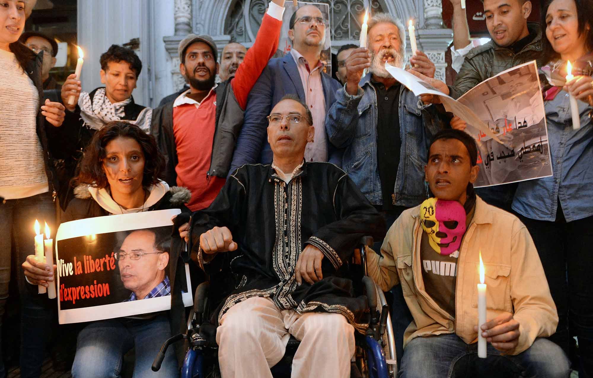 L'immagine mostra un gruppo di persone radunate in un ambiente urbano, apparentemente in un momento di protesta o di vigilanza. Alcuni partecipanti tengono in mano candele accese, mentre altri mostrano cartelli e manifesti con slogan che richiamano il tema della libertà di espressione. Tra le persone c'è un uomo in sedia a rotelle, che sembra essere al centro dell'attenzione. L'atmosfera è di solidarietà e impegno per i diritti civili.
