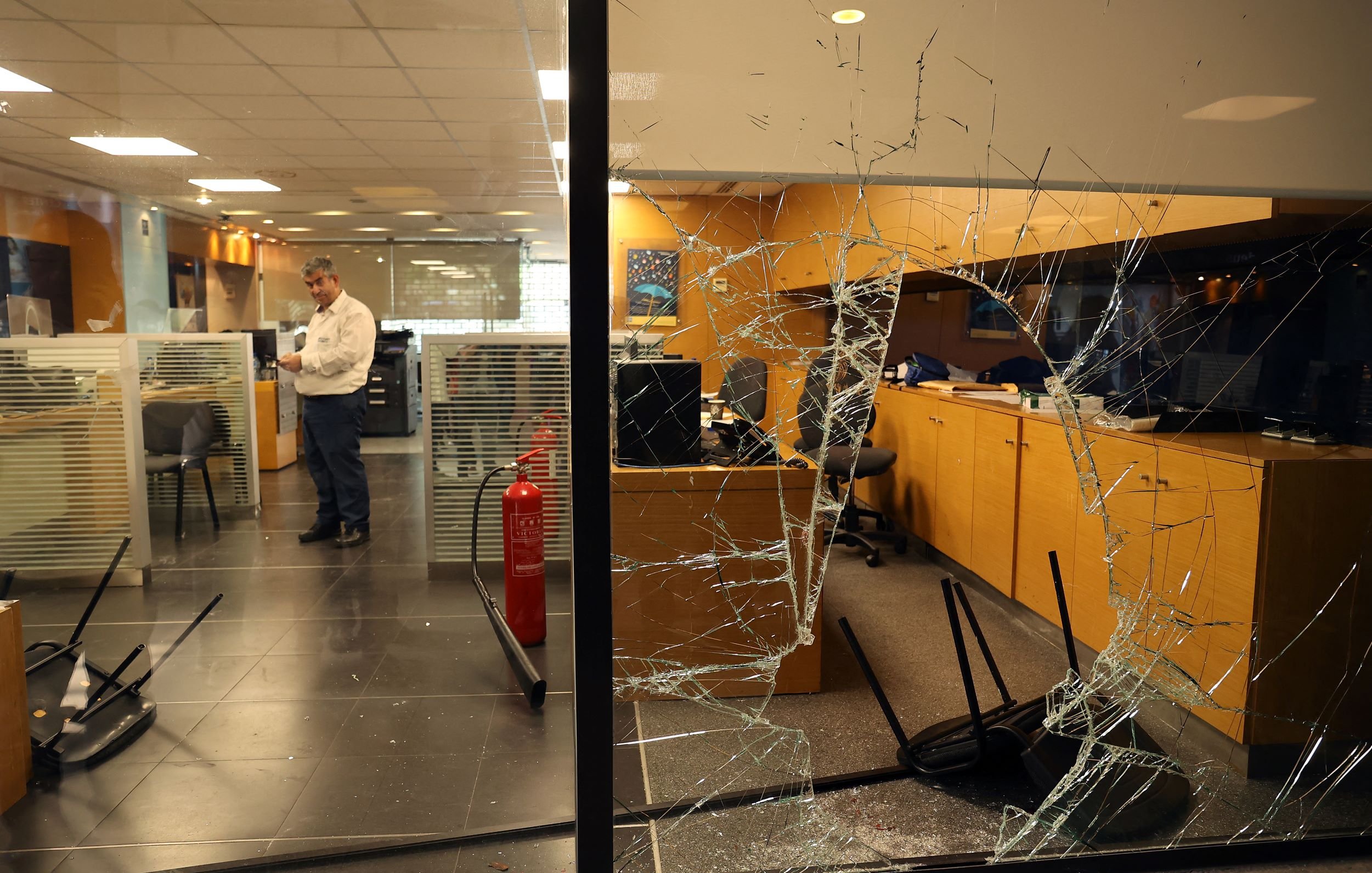 این تصویر یک نمای داخلی از یک دفترکار را نشان می‌دهد که به وضوح آسیب دیده است. شیشه‌ی پنجره شکسته و تکه‌های آن روی زمین پخش شده‌اند. در داخل اتاق چند صندلی معکوس و به هم ریخته وجود دارد. یک مرد در حال ایستادن در نزدیکی کامپیوترها به نظر می‌رسد و مشغول بررسی وضعیت است. همچنین در گوشه‌ای از اتاق، یک کپسول آتش‌نشانی قرار دارد. فضای کلی تصویر حاکی از بی‌نظمی و آسیب‌های شدید در این محیط کاری است.