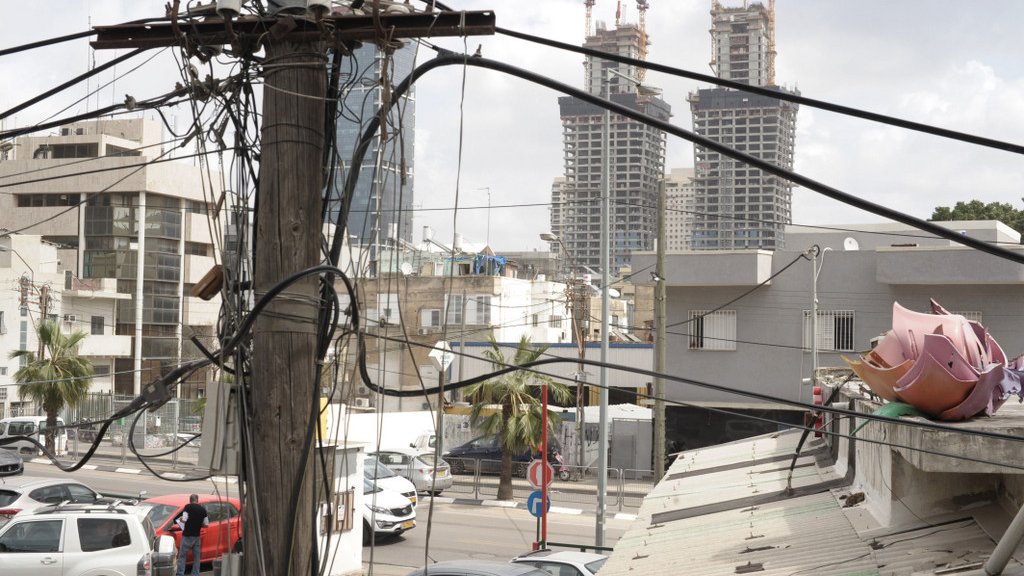 L'image montre une vue urbaine où plusieurs poteaux électriques sont entremêlés de fils. À l'arrière-plan, on peut apercevoir deux grands immeubles en construction. Au premier plan, il y a des bâtiments plus petits et des véhicules stationnés. On peut également voir des palmiers, ce qui indique un environnement peut-être chaud ou méditerranéen. L'ensemble donne une impression de ville en développement, avec un mélange de structures anciennes et modernes.