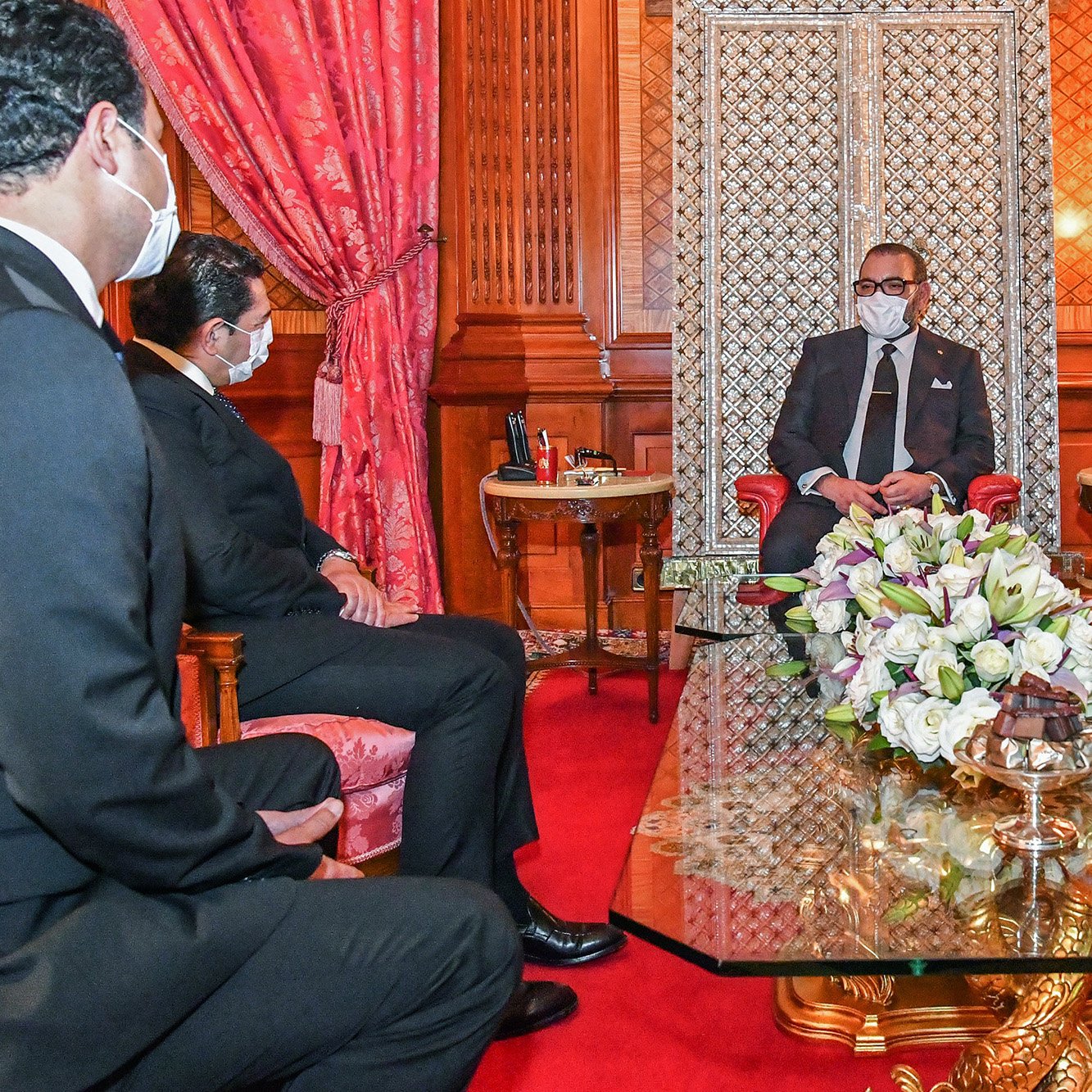 در این تصویر، یک جلسه رسمی در یک اتاق مجلل نمایش داده شده است. در مرکز تصویر، یک مرد (که به نظر می‌رسد مقام بلندپایه‌ای است) بر روی صندلی نشسته و با لباسی رسمی و ماسک به چهره دارد. سه مرد دیگر در سمت چپ و راست او نشسته‌اند و به او گوش می‌دهند. فضای اتاق با فرش قرمز و دیوارهای چوبی تزئین شده و نور ملایمی از چراغ‌های شمعی درخشان است. بر روی میز مقابل آنها دسته‌ای از گل‌های زیبا قرار دارد.