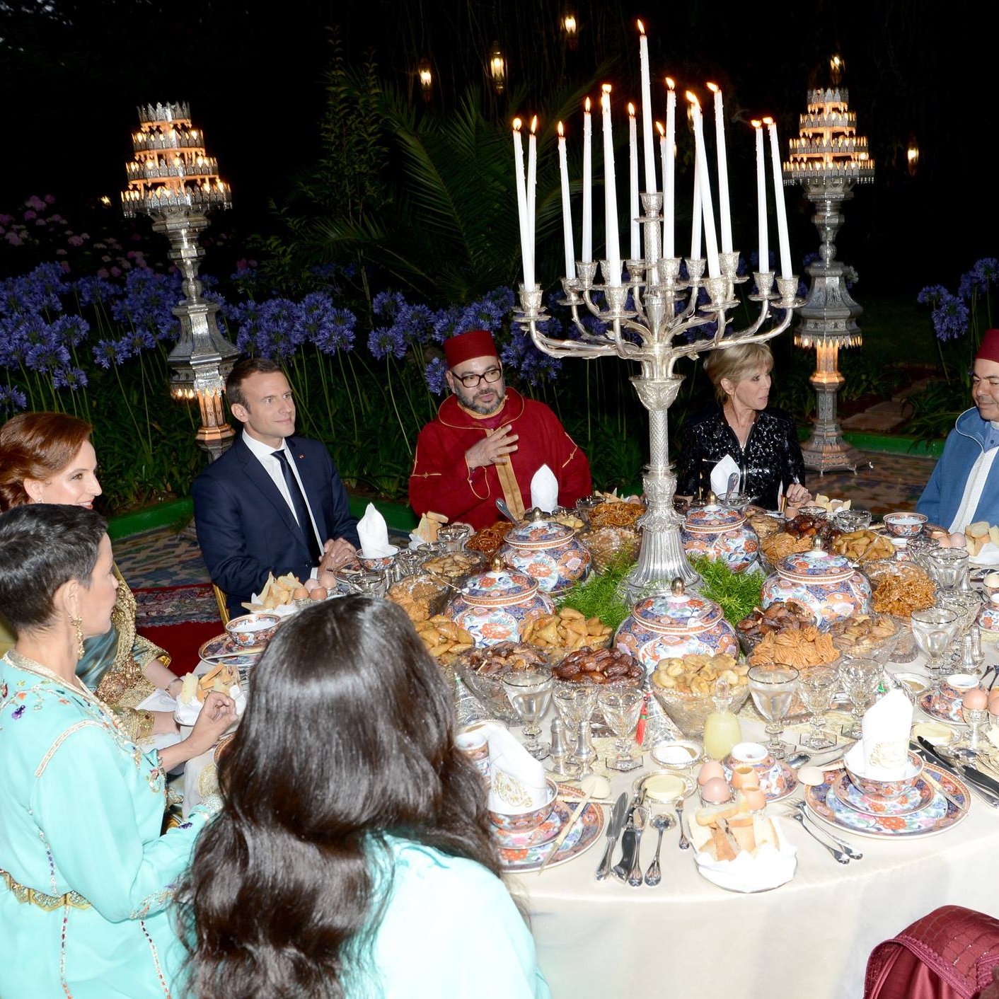 L'image montre une scène de dîner élégant, probablement dans un cadre extérieur. Une grande table est dressée avec une variété de plats et de délicieuses spécialités culinaires. De nombreuses bougies sont allumées, créant une ambiance chaleureuse et festive. Des convives, habillés en vêtements traditionnels, sont assis autour de la table, engageant des discussions animées. Au fond, on peut apercevoir des éléments décoratifs et des personnes en costume qui ajoutent à l'atmosphère formelle de l'événement.