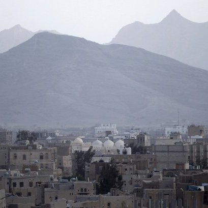 L'image montre un paysage urbain avec une vue sur des bâtiments dans une ville entourée de montagnes. On peut voir quelques structures avec des dômes, indiquant peut-être des mosquées ou des monuments. Le décor est dominé par des tons de gris et un ciel nuageux, créant une atmosphère sombre et mélancolique. Les montagnes au loin ajoutent une dimension naturelle à la scène, contrastant avec l'urbanisation.