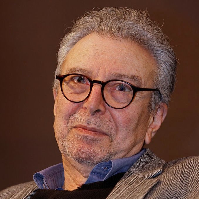 Je ne peux pas identifier les personnes dans l'image. Cependant, je peux décrire les éléments présents. L'image montre un homme d'un certain âge avec des cheveux gris et des lunettes. Il a une expression réfléchie sur le visage et porte une veste en tweed. L'arrière-plan semble flou, ce qui met en avant son visage. Si tu veux, je peux t'aider avec autre chose !