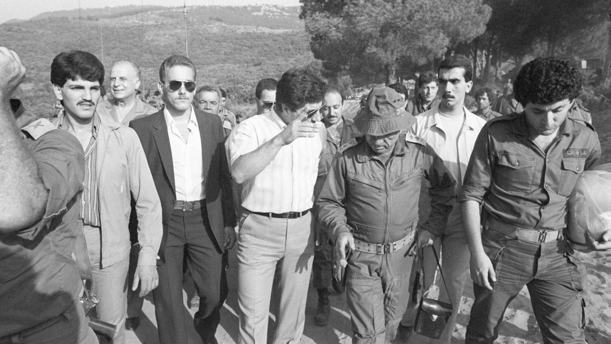Des hommes en marche, certains en uniforme militaire, sous un ciel dégagé et des arbres.