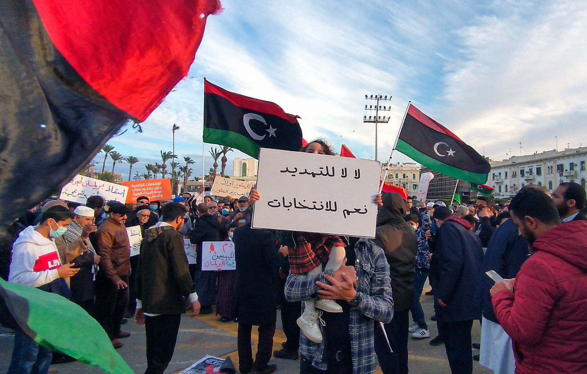 L'image montre une manifestation avec de nombreuses personnes rassemblées. Des drapeaux de la Libye flottent dans l'air, affichant des couleurs rouge, noir et vert. Au centre de la scène, une personne tient une pancarte avec des inscriptions en arabe. On peut également voir des participants portant des masques et d'autres pancartes, exprimant des revendications politiques. L'ambiance paraît dynamique, avec une multitude de personnes engagées dans une cause commune.