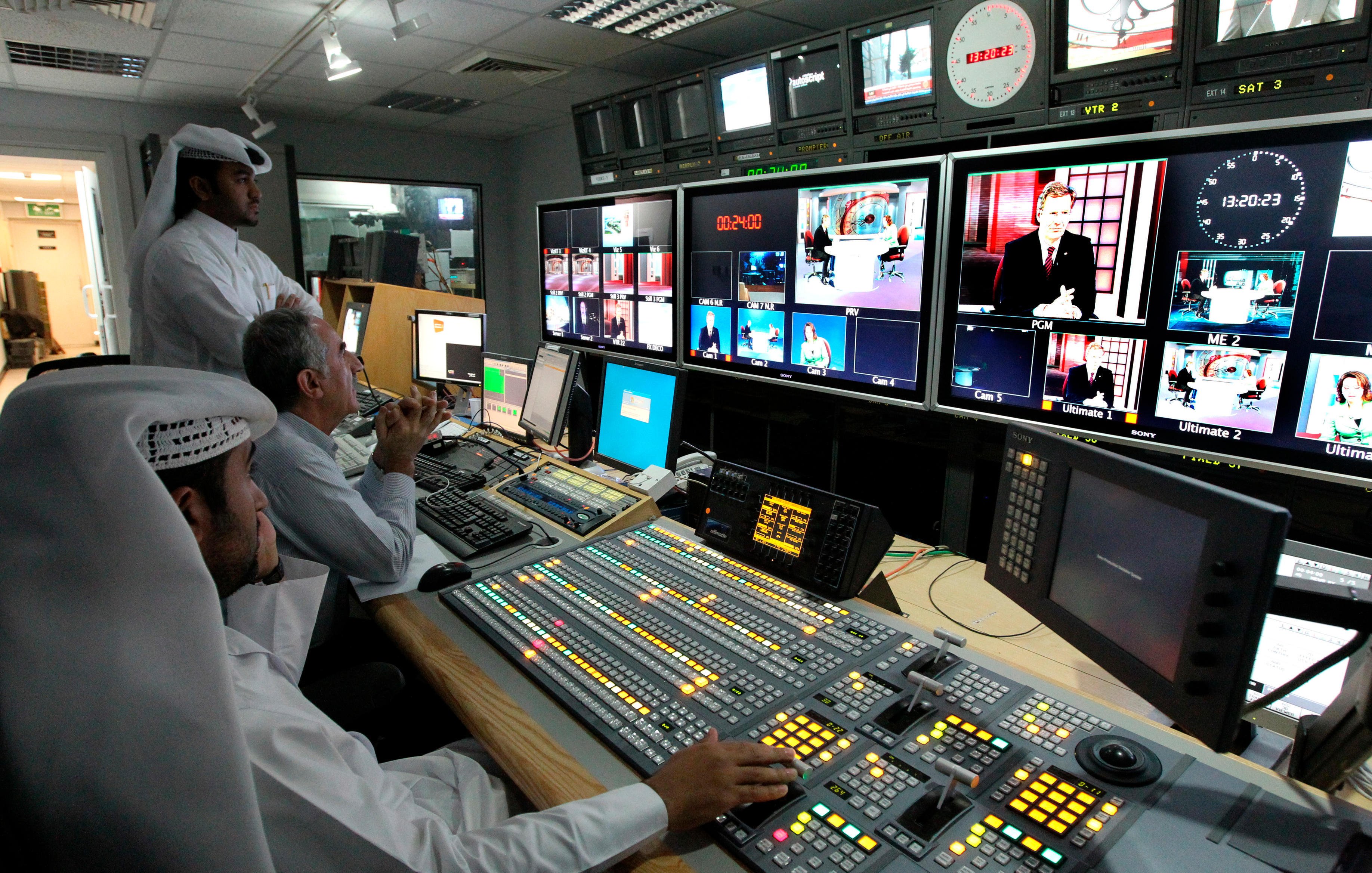 تظهر الصورة غرفة تحكم تلفزيونية. في المقدمة، يوجد شخصان يجلسان أمام معدات التحكم، مع وجود مجموعة من الأزرار والشاشات! أحدهم يبدو مركزًا على العمل، بينما الآخر يحمل هاتفًا ويبدو مشغولًا. في الخلف، يقف شخص ثالث يتابع الشاشات. تبرز من خلفهم مجموعة من الشاشات الكبيرة تعرض برامج وأخبار مختلفة، مع وجود ساعة تُظهر الوقت. الإضاءة في المكان طبيعية وتضفي أجواء احترافية.