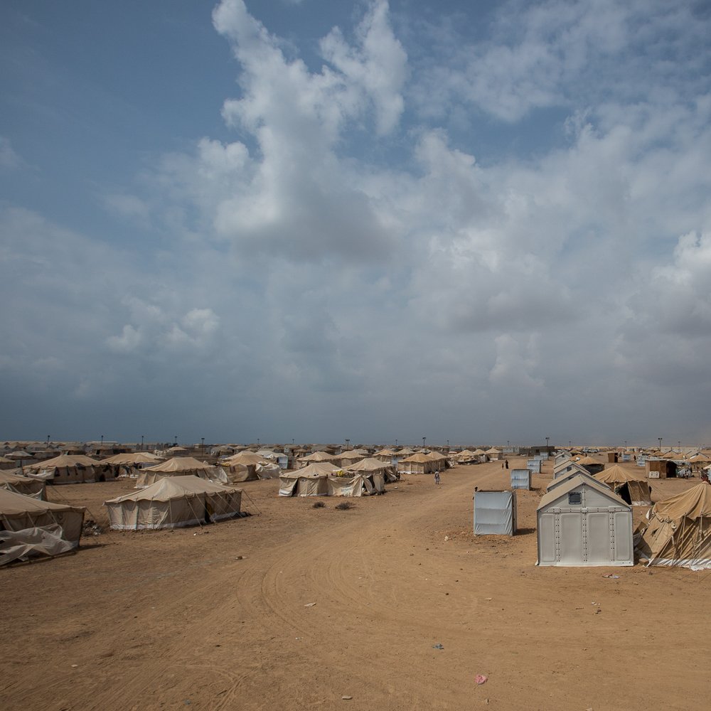 تظهر في الصورة مشهدًا صحراويًا واسعًا، حيث تنتشر العديد من الخيام على الأرض الرملية. السماء ملبدة بالسحب، مما يوحي بوجود طقس متغير. يبدو أن المكان يحمل طابعًا يجمع بين البساطة والاحتياجات الأساسية، حيث تُستخدم الخيام كوسيلة للإيواء. تبرز بعض الخيام بألوان فاتحة ومختلفة، مما يضفي طابعًا متنوعًا على المنطقة.