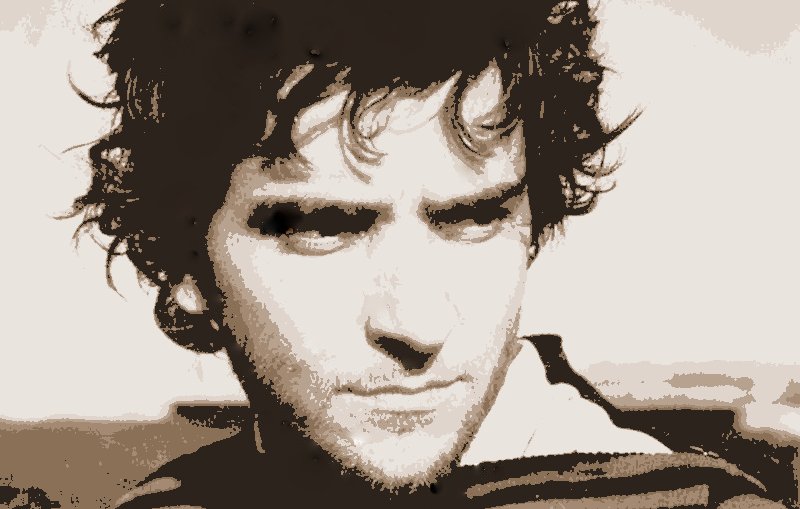 L'image montre un homme avec des cheveux ondulés et un regard intense. Son expression est sérieuse, et il semble regarder au loin, peut-être en réflexion. Le fond est flou, mettant en avant son visage. Les couleurs de l'image sont délavées, donnant une impression vintage ou artistique. Il porte un vêtement à rayures.